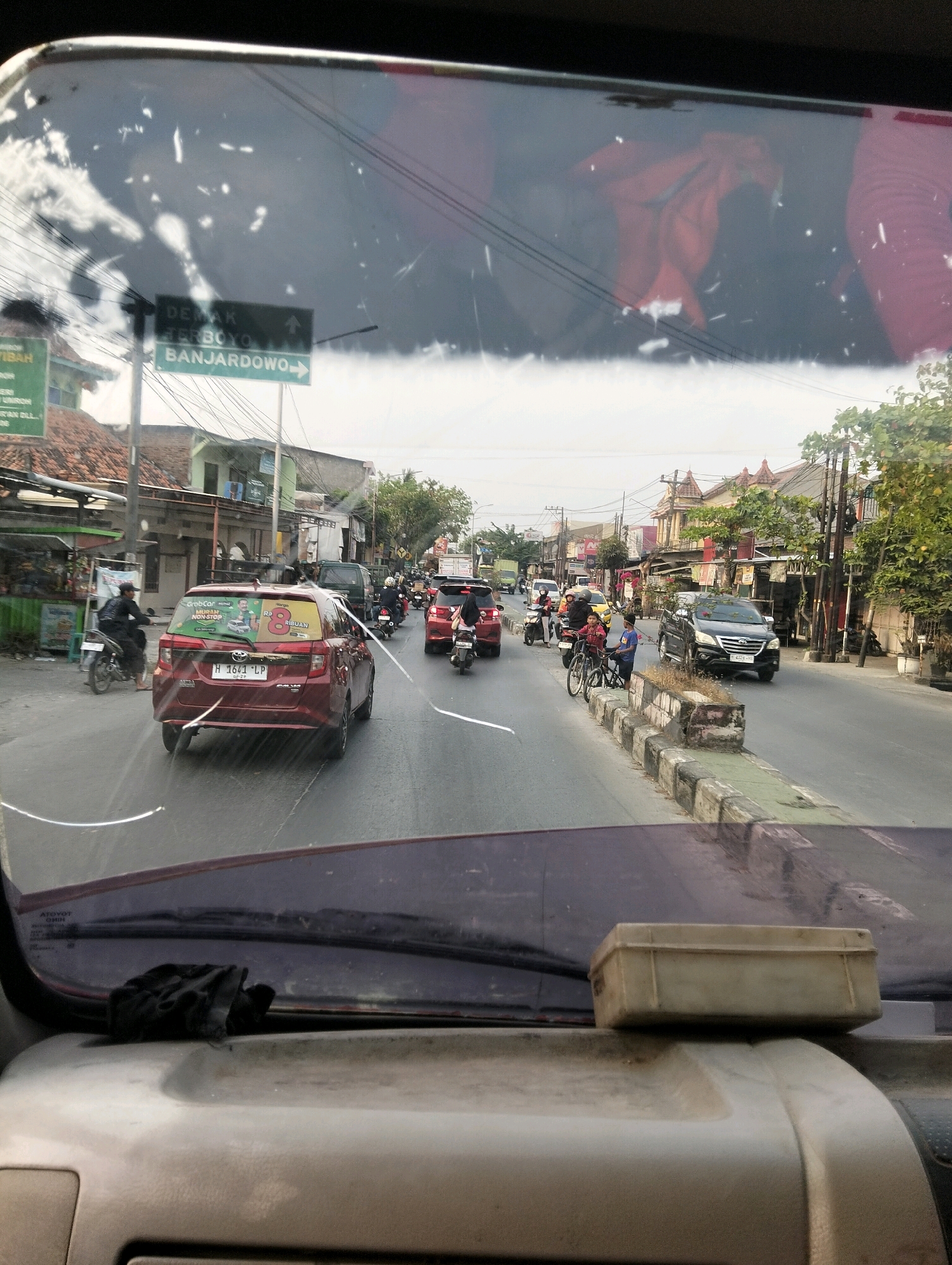 The image shows a street scene in Semarang, Indonesia, viewed from the interior of a vehicle. The foreground is dominated by the dashboard and the lower portion of the windshield. In the midground, traffic is moving along a paved road lined with buildings and trees. A red car with license plate H 1641 LP is prominently in the center, with a sticker on its rear window that includes a Grab logo and the number "8". Other vehicles, including motorcycles and cars, are visible in the traffic flow. A directional signpost indicates "DEMAK TERBOYO BANJARDOWO". The environment appears to be urban with residential and commercial buildings. Reflections on the windshield obscure some details, and there are cracks visible on the glass.