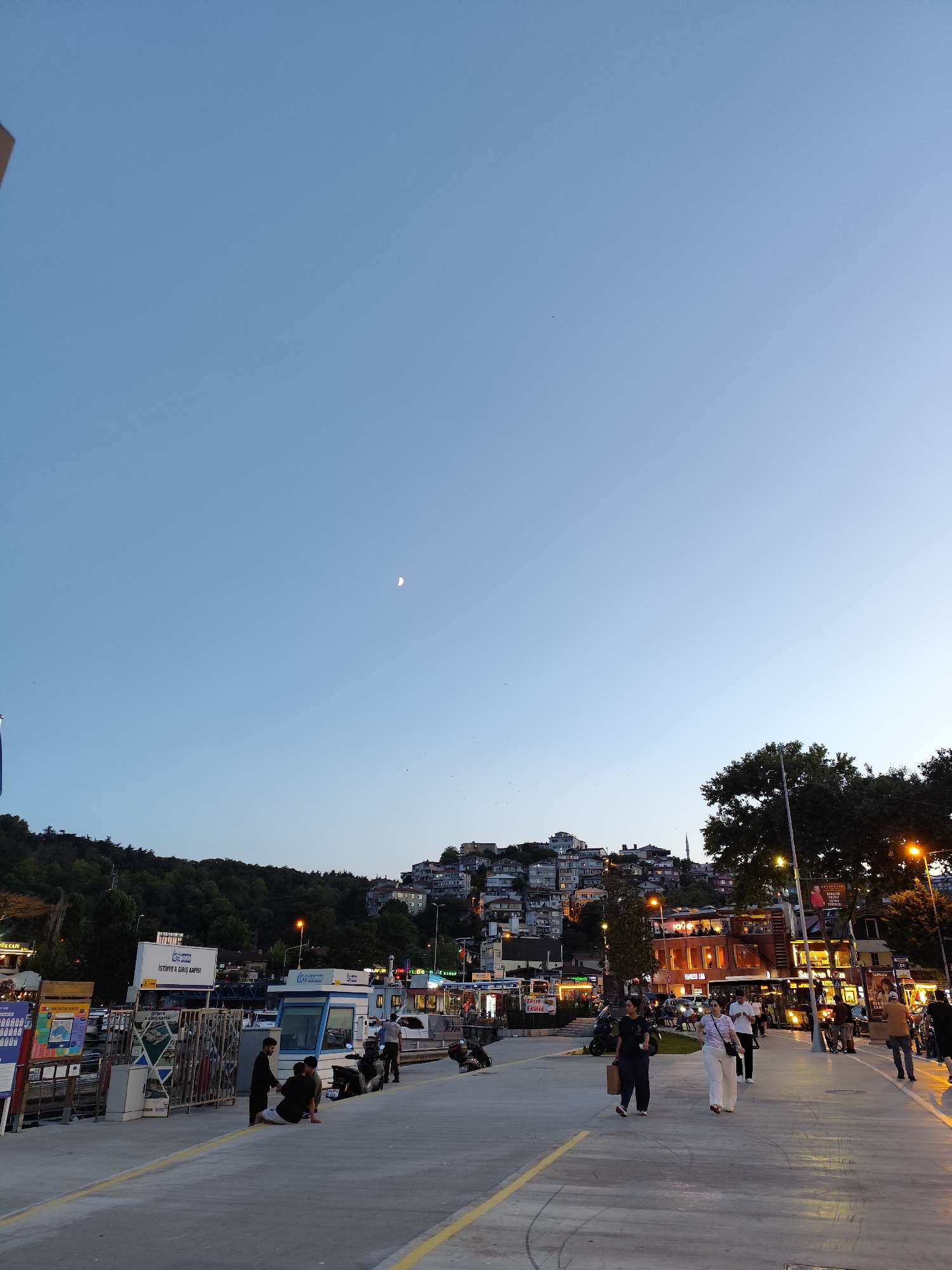 The image captures a twilight scene on a paved waterfront promenade in Sarıyer, Turkey, overlooking a hillside densely packed with residential buildings. The sky is a gradient of clear blue to a lighter hue near the horizon, with a sliver of the moon visible. A tree-lined slope rises behind the promenade, topped by the cluster of houses.
On the promenade, several individuals are present. In the foreground, a group of young men are interacting, with one sitting on the ground and two standing. Further along the promenade, people are walking, including two women in the mid-ground, one carrying a bag. The promenade is marked with yellow lines.
Various structures are situated along the promenade. To the left, there are kiosks and signs, including one with blue and white branding that reads "İSTİNYE A GİRİŞ KAPISI" (Istinye Entrance Gate). Further to the right, there are more commercial establishments, some illuminated with artificial lights, suggesting shops and cafes. Streetlights are also visible, illuminating sections of the promenade. The scene appears to be a public space, possibly a ferry terminal or a recreational area, given the presence of kiosks and the waterfront setting. The overall atmosphere is calm, transitioning from daylight to evening.