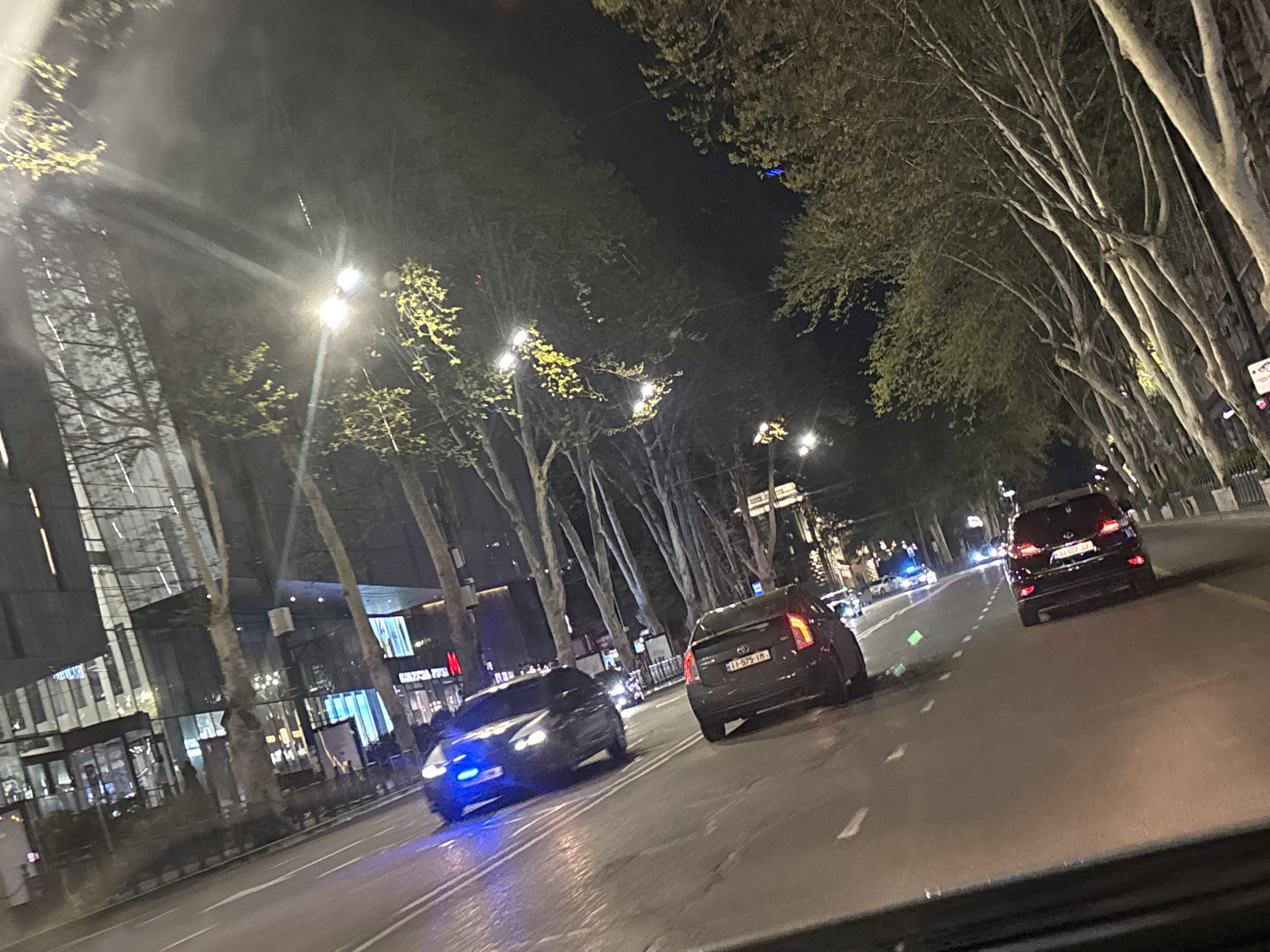 The image captures a nighttime street scene in an urban environment, likely from the perspective of a vehicle. The setting is a multi-lane road flanked by tall, slender trees with some budding light green leaves, illuminated by numerous streetlights that cast bright glows and lens flares. Modern glass-fronted buildings are visible on the left side of the road, suggesting a commercial or mixed-use area.
Several cars are present on the road. In the foreground on the left, a dark-colored sedan, possibly a Toyota Prius, is moving with prominent blue flashing lights at its front. Its license plate appears to be "II-575-IM". On the right, further down the road, a dark SUV, potentially a Hyundai Santa Fe, displays illuminated taillights. Its Georgian license plate clearly reads "JJ-027-JJ". In the distance, other vehicle lights are visible, including what appears to be another white car with blue flashing lights, possibly an emergency or special service vehicle.
The activity is typical nighttime traffic flow on a city street. The time of day is unequivocally night, as indicated by the darkness and the reliance on artificial lighting from streetlights and vehicle head/taillights. There are no clear indications of people or specific weather conditions other than a clear night.
Visible text includes the Georgian license plates on the vehicles: "II-575-IM" on the sedan and "JJ-027-JJ" on the SUV. Faint signage for "H&M" can be discerned on one of the commercial buildings in the background. The overall impression is a bustling yet dimly lit city thoroughfare.
