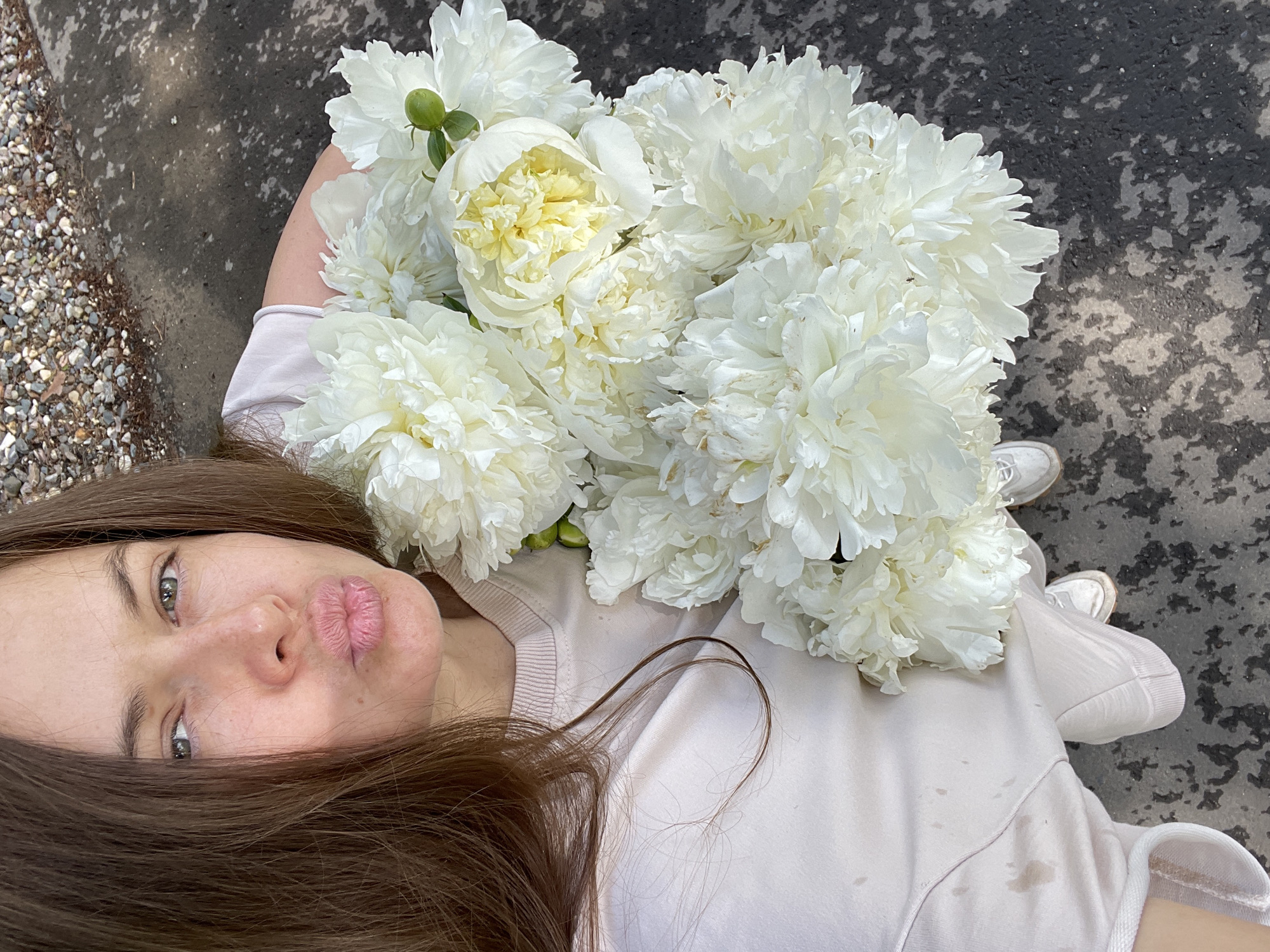 A woman with long, brown hair, is taking a selfie. She is wearing a light pink sweatshirt and light pink sweatpants. Her lips are pursed in a kiss. She is holding a large bouquet of white peonies. The background appears to be an outdoor setting, possibly a street or sidewalk, with some gravel and asphalt visible. The lighting suggests it is daytime. The location is identified as Zarechny, Russia.