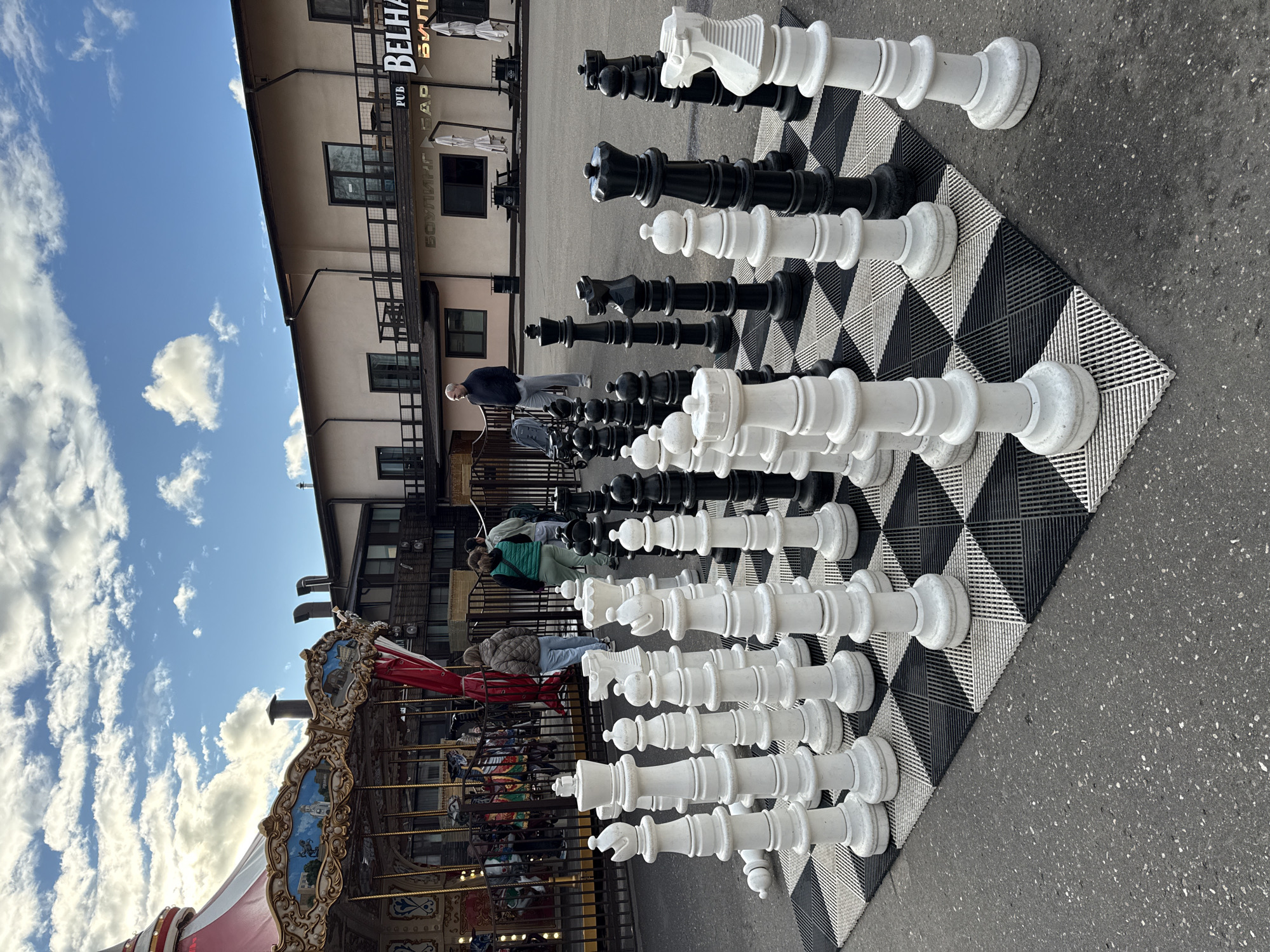 The image shows a giant chess set outdoors in Peresvet, Russia. The chess pieces are white and black, positioned on a large checkerboard mat on a paved area. In the background, there is a building with a sign "BOULING PUB BELHA БУЛИНГ". To the left of the chess set, there's a carousel with various painted scenes. Several people are present near the chess set and carousel. The sky is blue with scattered white clouds.