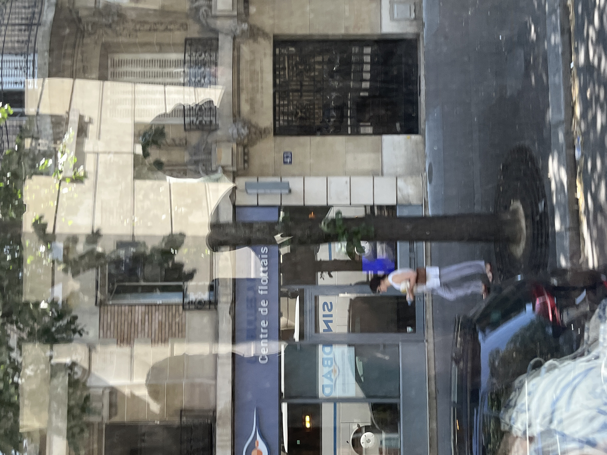 The image shows a street scene in Paris, France, with a focus on the facade of a building and the street in front of it. The building has a light-colored stone facade with architectural details like balconies and shutters. A sign on the building reads "Centre de flottais". A woman in a white top and grey pants is walking on the sidewalk, holding a small object in her hand. A dark-colored car is parked on the street, partially visible in the foreground. The scene is dappled with light and shadow from trees overhead, indicating it is daytime. The street surface is asphalt, with a visible curb and sidewalk. Reflections are visible on the building's windows, suggesting the image was taken from across the street or from a moving vehicle.