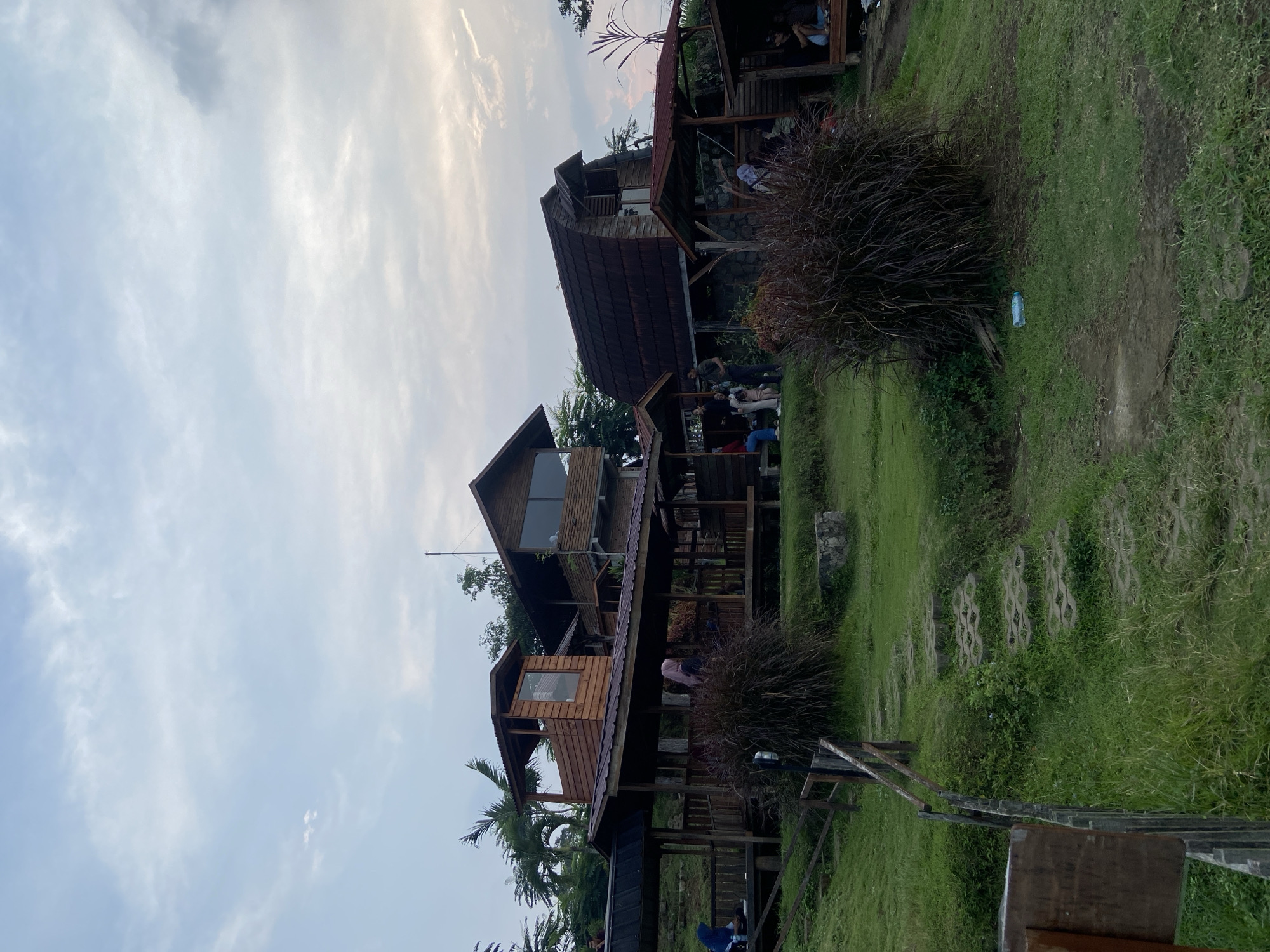 The image depicts a rural setting in Selotapak, Indonesia, featuring wooden structures resembling multi-level huts. The huts appear to be a part of a gathering place, with people sitting and socializing inside and around the structures. The foreground includes lush green grass and foliage, with stepping stones creating a pathway. The sky is overcast with a muted color palette.