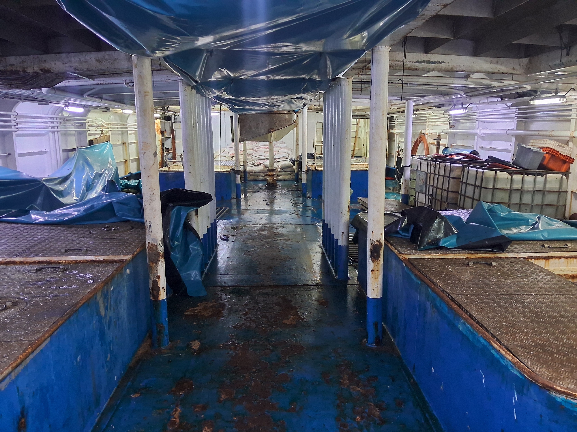 The image depicts the interior of a cargo ship's hold in Chimbote, Peru. The space is rectangular with a blue floor that shows signs of wear and tear, including rust and water stains. Metal columns painted white with blue bases support a metal ceiling structure covered in blue tarps. The sides of the hold are lined with blue metal structures with diamond-plated tops, also showing rust. Various objects are present, including stacks of white sacks in the background, large plastic containers, orange crates, and a life preserver. Blue tarps cover portions of the hold, suggesting storage or protection of goods. The lighting appears to be artificial, coming from overhead fixtures. The overall impression is one of a utilitarian and somewhat weathered industrial environment.