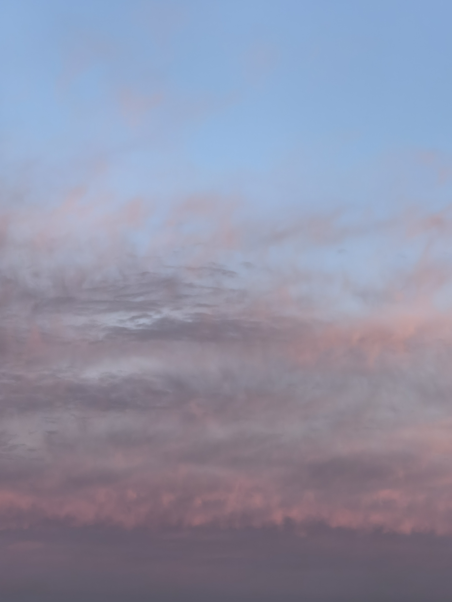 The image displays a sky with a gradient of colors, transitioning from a pale blue at the top to hues of pink and purple towards the bottom. Wispy clouds are scattered throughout, with some tinged with pink, suggesting either sunrise or sunset. The lower portion of the image is darker, with the pink and purple tones becoming more dominant, indicating the presence of thicker cloud cover or a deeper part of the sky. The scene is devoid of any terrestrial elements or identifiable structures, focusing solely on the atmospheric conditions. The location is described as Unknown, Unknown.