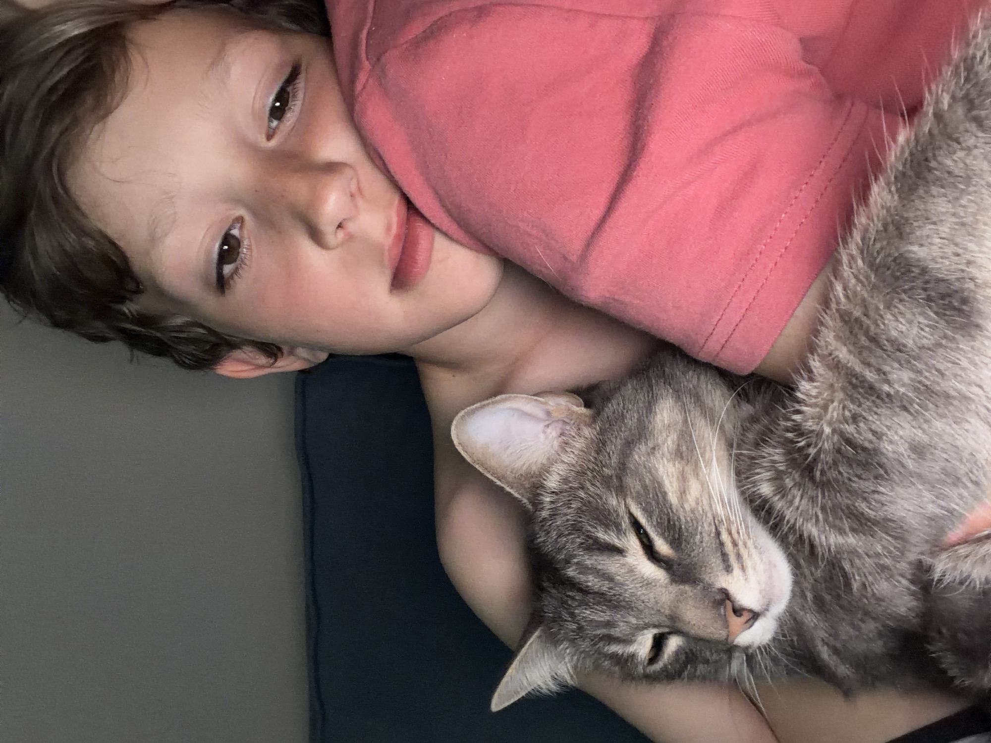 A boy with brown hair and eyes is lying on his side, his head resting on a pink t-shirt. He is cuddling with a gray tabby cat, which is also lying on its side with its eyes mostly closed. The boy's arm appears to be around the cat. The background is a light gray and dark blue. The location is not discernible from this image and there is no mention of Chaikovsky, Russia.