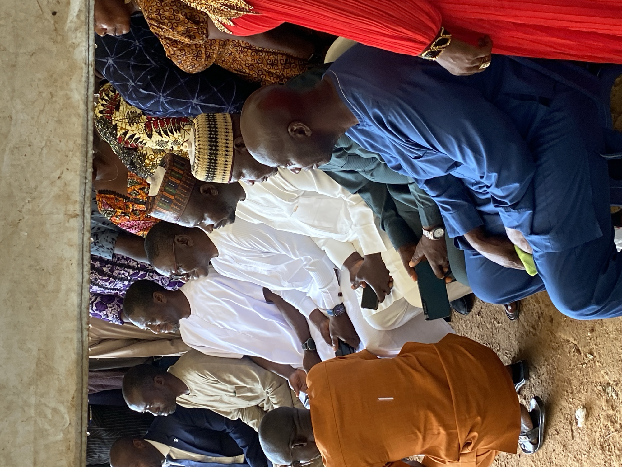 In Kuru, Nigeria, a group of men and women are gathered, seated and observing an event. The setting appears to be outdoors, with a dirt ground surface and a simple overhead structure that casts a shadow. Several men are dressed in traditional attire, including long, flowing white robes and embroidered caps. One man in the foreground wears a vibrant blue traditional outfit, while another is dressed in a bright orange ensemble and is crouching. Women are visible in the background and to the right, adorned in colorful patterned fabrics and headwraps. Some individuals are holding or looking at their mobile phones. The overall atmosphere suggests a formal gathering or ceremony.