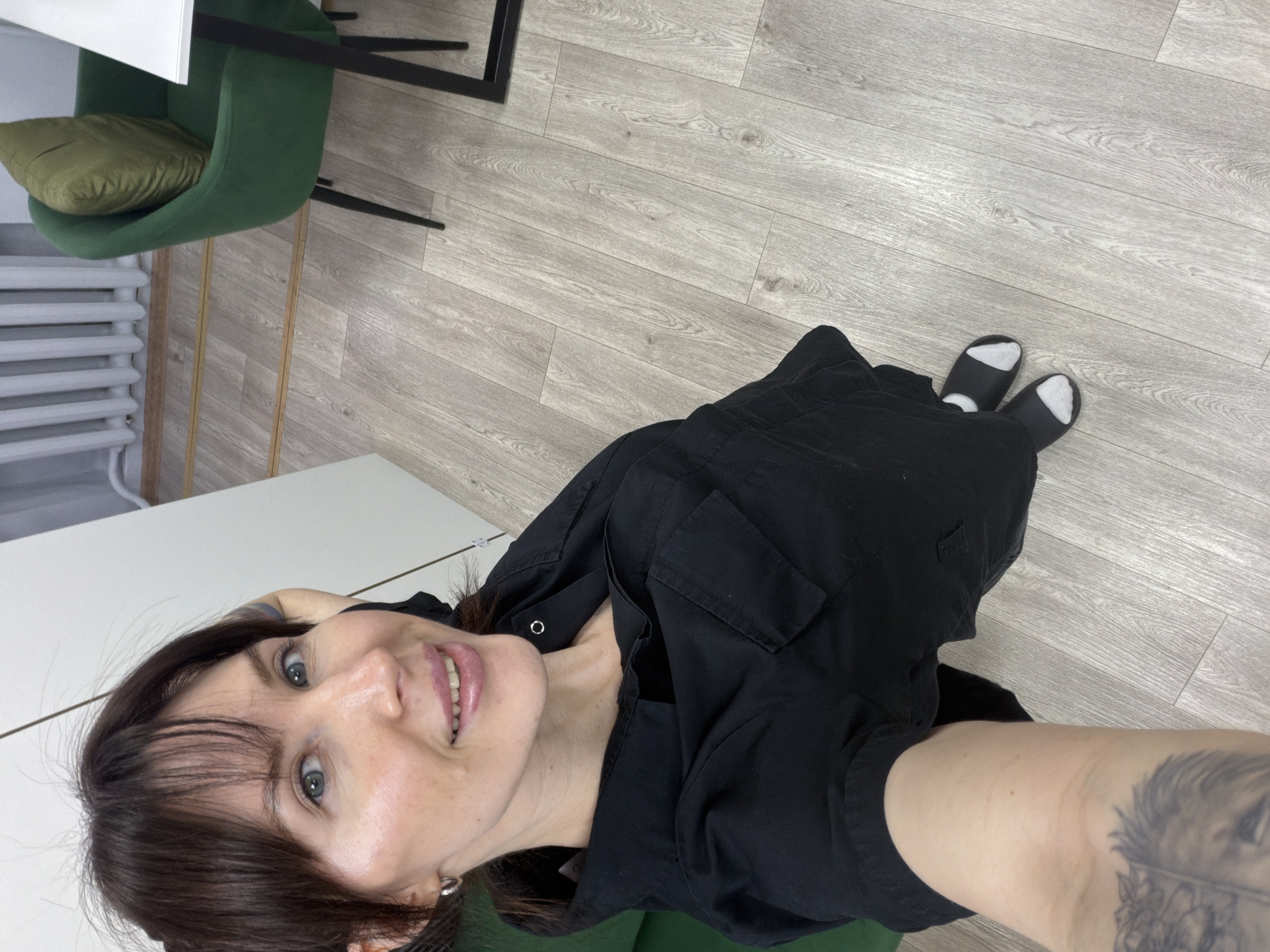 A woman with dark hair and fair skin is taking a selfie in what appears to be an indoor office or studio setting. She is wearing a black shirt and dark socks. A green chair with a pillow is located near a white table. The flooring is light-colored wood-look laminate. A radiator can be seen in the background. This image was found online and identified as being taken in Perm, Russia.