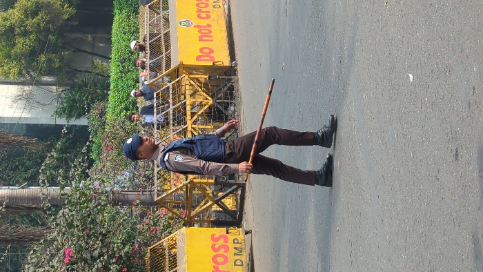 A uniformed police officer, wearing a blue cap and vest over a tan shirt, stands on an asphalt road holding a long stick. Behind him is a yellow barrier with "Do not cross" printed on it, partially obscured by foliage and a metal fence. Other individuals stand behind the fence. The image is slightly blurred, suggesting it was taken at a distance.