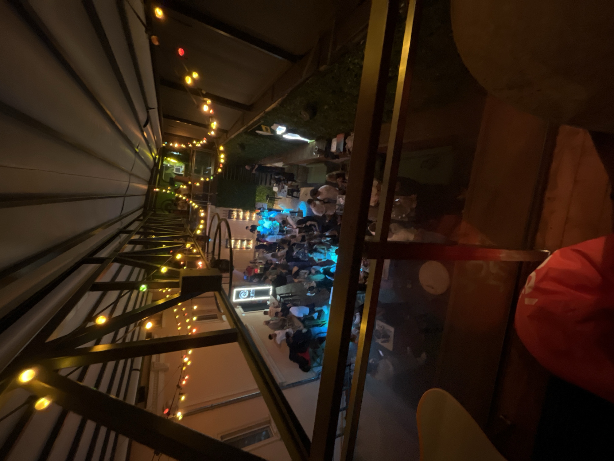 The image shows a dimly lit interior space with a balcony overlooking a crowded area. On the balcony, there are dark metal beams and strings of yellow and red lights. Below, in a multi-level area, many people are gathered, with blue and white lights illuminating the scene. Some individuals are standing and interacting, while others appear to be seated. The overall atmosphere suggests a social gathering or event. The location is identified as the "Apothecary" club in Plovdiv, Bulgaria.