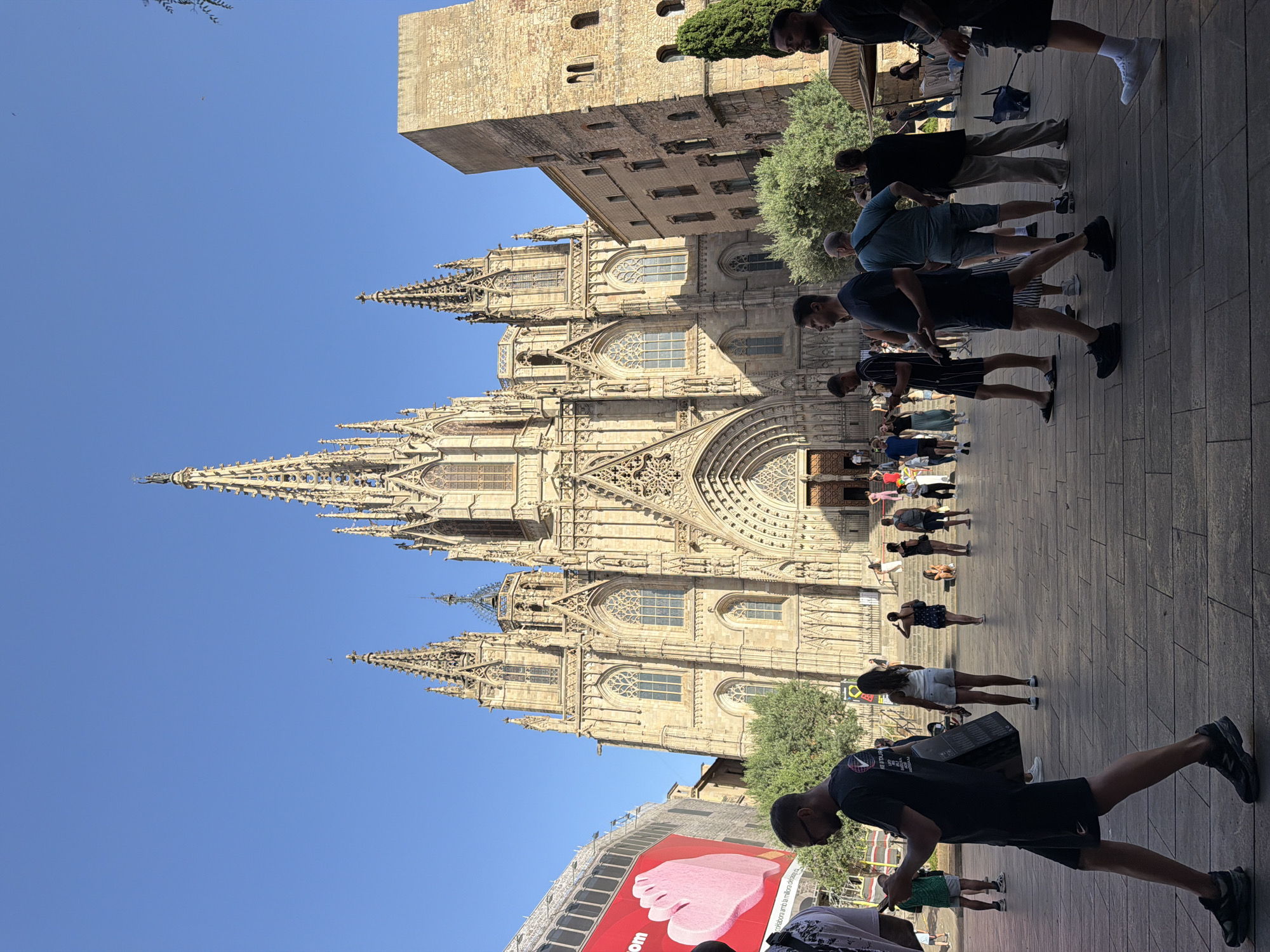 This is a photograph of the Barcelona Cathedral (Catedral de Barcelona) in the Gothic Quarter of Barcelona, Spain. The image is taken from a low angle, looking up at the cathedral's facade, which is ornate and features Gothic architecture with spires and arched windows. In the foreground, a paved plaza is occupied by numerous people walking and standing.
On the left side of the image, a large red billboard with an image of a pink hand is visible, along with part of a modern building. The cathedral's facade dominates the background. To the right of the cathedral, a tall, stone tower, possibly part of an older structure, stands prominently.
The people in the foreground are diverse in age and attire, many dressed in casual summer clothing. Some are walking, some are conversing, and others are looking at their phones. The sky is clear and blue, indicating a sunny day. The overall scene depicts a lively public space in front of a significant historical landmark.