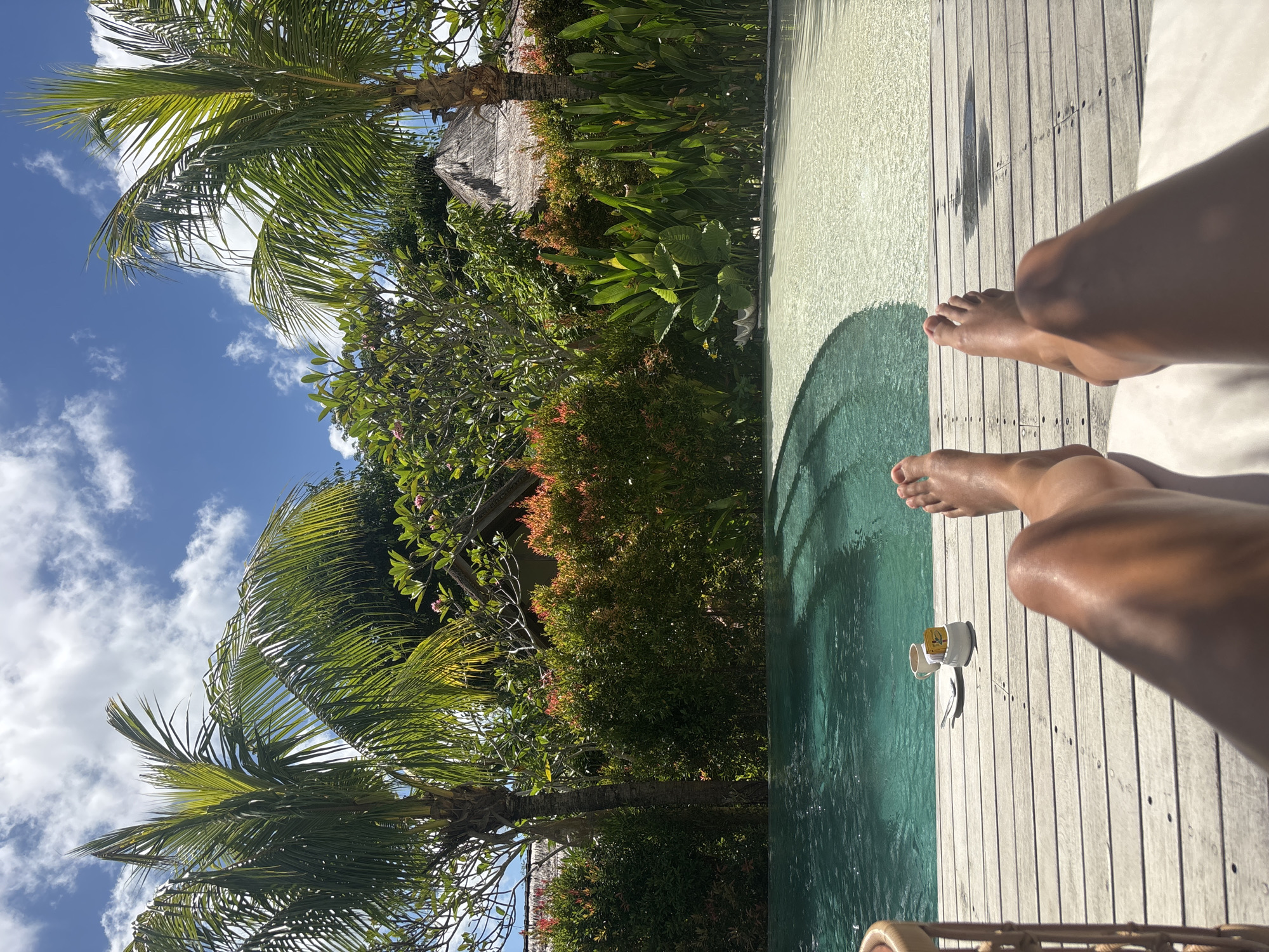 A first-person view, rotated 90 degrees counterclockwise, shows tanned human legs and feet resting on a light-colored wooden deck adjacent to a swimming pool. The pool's edge is visible on the left side of the frame, transitioning from a clear, light blue to a darker green where there is more depth. Next to the pool and deck is lush vegetation, including trees, bushes, and palm trees. A small structure with a thatched roof is partially visible amidst the foliage. The sky is blue with scattered clouds. A small white cup and saucer are on the deck near the pool's edge. The scene appears to be in Kuta Selatan, Indonesia.