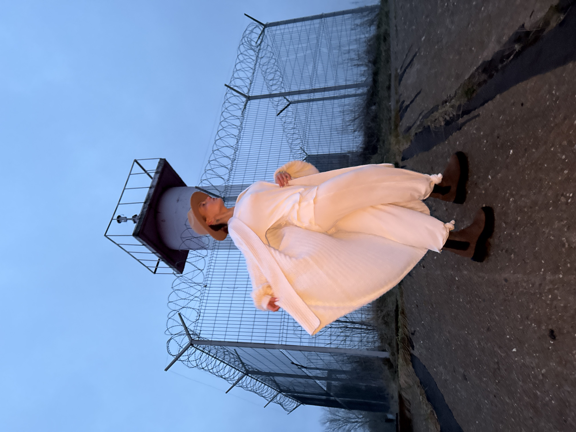 The image captures a full-body portrait of a person standing outdoors in what appears to be a secure or industrial setting. The lighting suggests either twilight or an overcast day, with a uniform pale blue-grey sky dominating the upper portion of the frame.
The central subject is a female, seen in profile, looking upwards and slightly to her left. She is dressed entirely in light-colored (off-white or cream) clothing, which includes a loose-fitting long-sleeved top, wide-leg pants, and a long, flowing cardigan or wrap. A light beige or brown wide-brimmed hat is on her head, and she wears dark brown ankle boots. Her hands are partially visible, one lightly resting on her clothing.
Behind the person, a tall metal fence topped with multiple strands of concertina wire (razor wire) stretches across the midground, suggesting a restricted area. To her left, partially obscured, is a dark, angular structure that resembles a watchtower or observation post, featuring a small platform at its apex with some equipment mounted. The foreground is a dark, uneven paved surface, likely asphalt or concrete, with scattered patches of dry grass and weeds.
The person is posed for the camera, appearing contemplative or observant of the sky or the tower. No active event is unfolding. There is no visible text in the image. The scene is set in an outdoor environment, consistent with a location like Chaikovsky, Russia, given the contextual information.