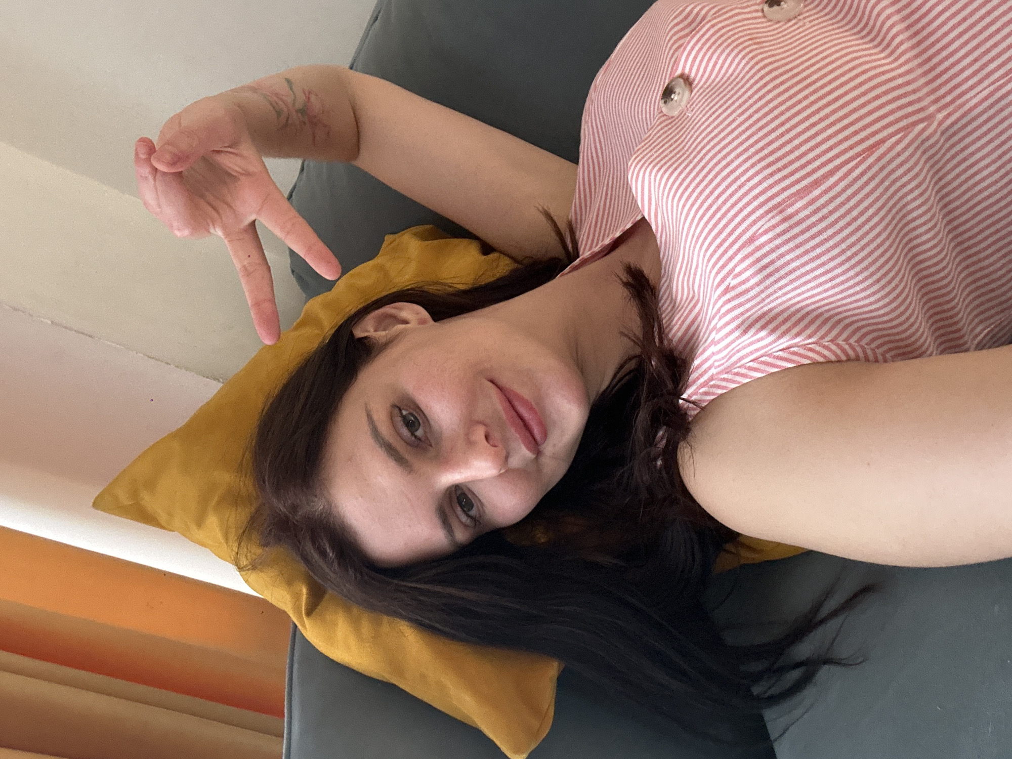 The image is a selfie of a woman lying on her side on a couch, propped up by a yellow pillow. She is wearing a pink and white striped shirt with two white buttons. Her arm is raised, and she is making a peace sign with her fingers. She has long, dark hair and is looking at the camera with a slight smile. The lighting suggests it is daytime, and the setting appears to be indoors, possibly a living room. There is a tattoo visible on her raised arm. The background includes a gray couch and some orange blinds.
