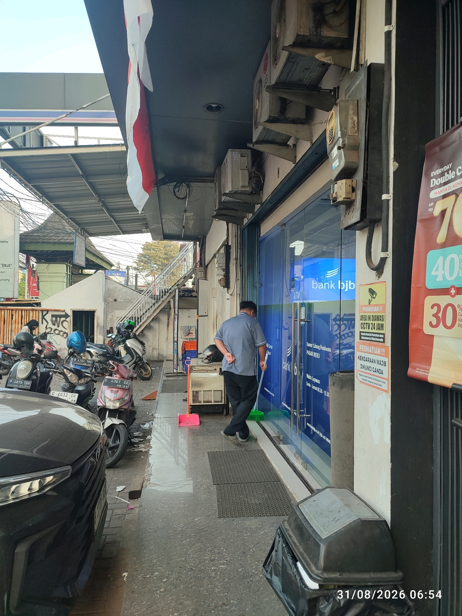 The image depicts an exterior view of a Bank BJB branch in East Jakarta, Indonesia, on August 31, 2026, at 6:54 AM. A man in a grey uniform is sweeping the sidewalk in front of the bank's glass entrance with a green broom. The bank's facade features large glass windows with the "bank bjb" logo visible. Several motorcycles are parked along the sidewalk, and a black car is partially visible on the left. Above the bank, an Indonesian flag hangs, partially obscured by an awning. Various air conditioning units are mounted on the exterior wall of the building. A large trash bin with a black plastic bag liner is situated near the corner of the building. Several warning signs are posted on the wall, including one indicating CCTV surveillance and another about mandatory double parking. In the background, a white staircase leads up to an upper level.