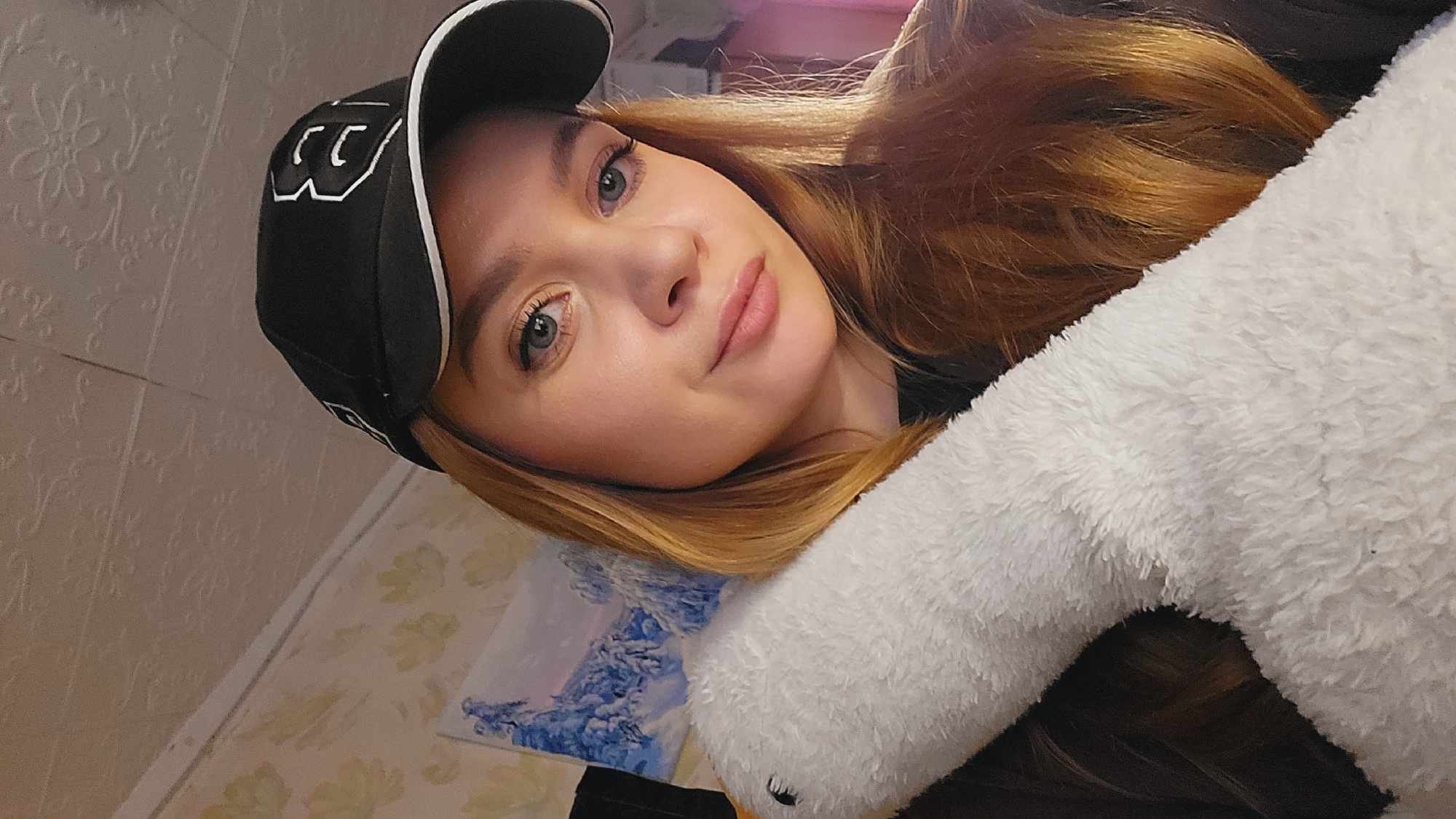 A woman with auburn hair is posing for a selfie, resting her head on a white, fluffy object, likely a stuffed animal or pillow. She is wearing a black baseball cap with white trim and a white "CC" logo. The background shows a wallpapered wall with a floral pattern, and a landscape painting or print with blue and white hues. The location, Зипуново, Russia, is not evident from the image.