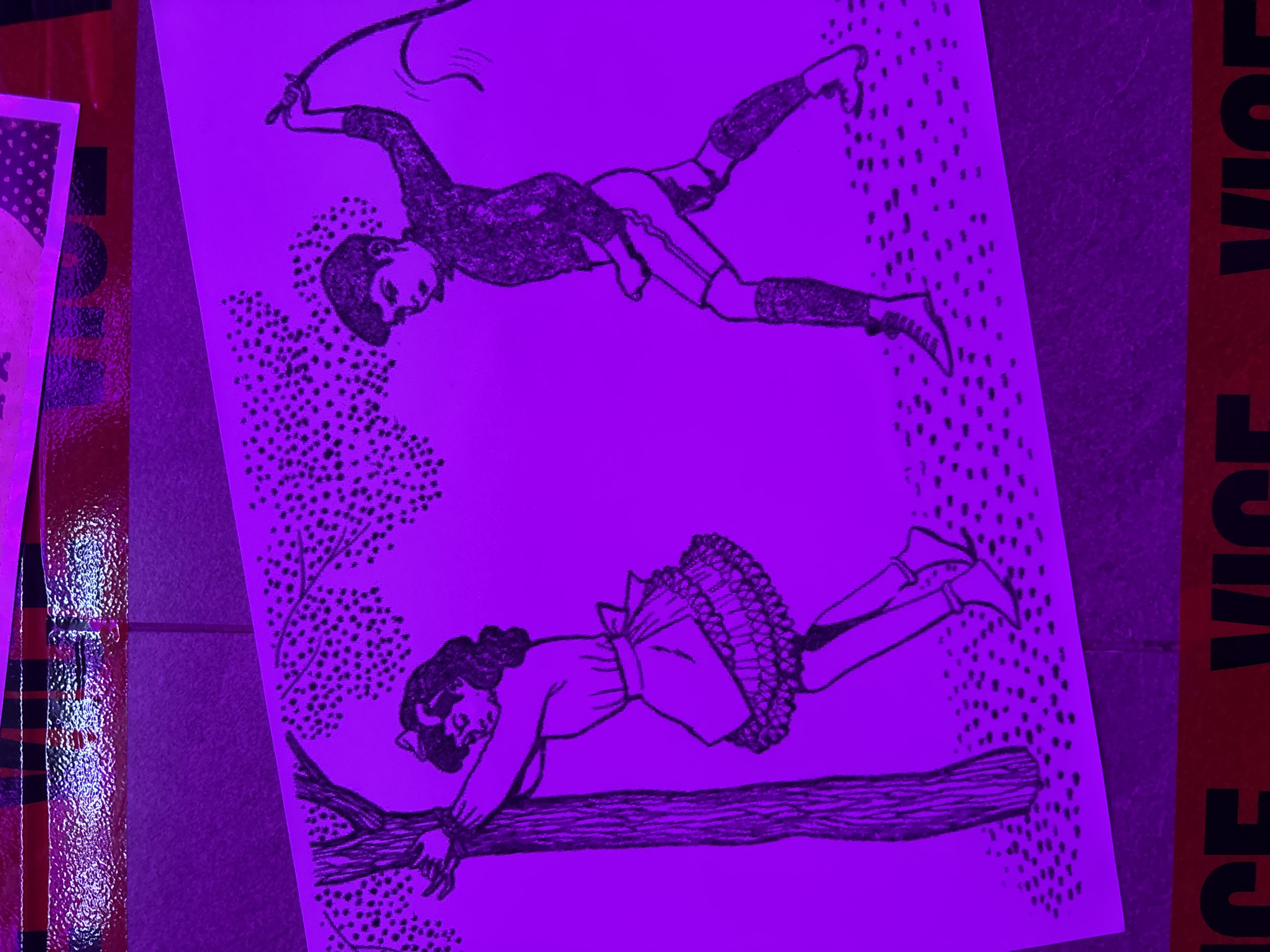 A black and white drawing of a girl tied to a tree and a boy holding a whip is displayed under purple lighting. The drawing is on a white sheet of paper, and the background behind it is blurred. In the foreground, a red and black striped tape is visible. The overall scene suggests a staged or artistic presentation rather than a natural environment.