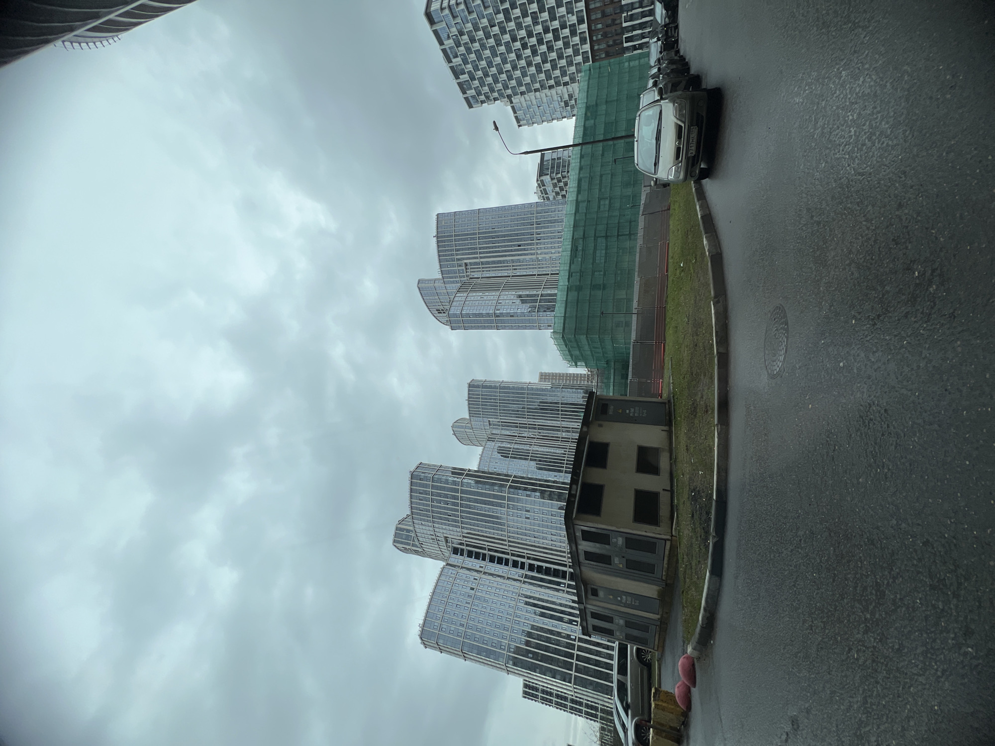 The image depicts a street scene in Khimki, Russia, under an overcast sky. Several modern, high-rise apartment buildings with mirrored glass facades are visible in the background. Construction appears to be underway on some of the buildings, with green netting draped on one side. A small, boxy, low-rise structure with multiple windows and doors sits at street level. The road is wet, suggesting recent rain. Several cars are parked along the curb, including a silver hatchback in the upper right. A street lamp is also visible. The environment suggests a modern urban area undergoing development. There are no people visible in the image.