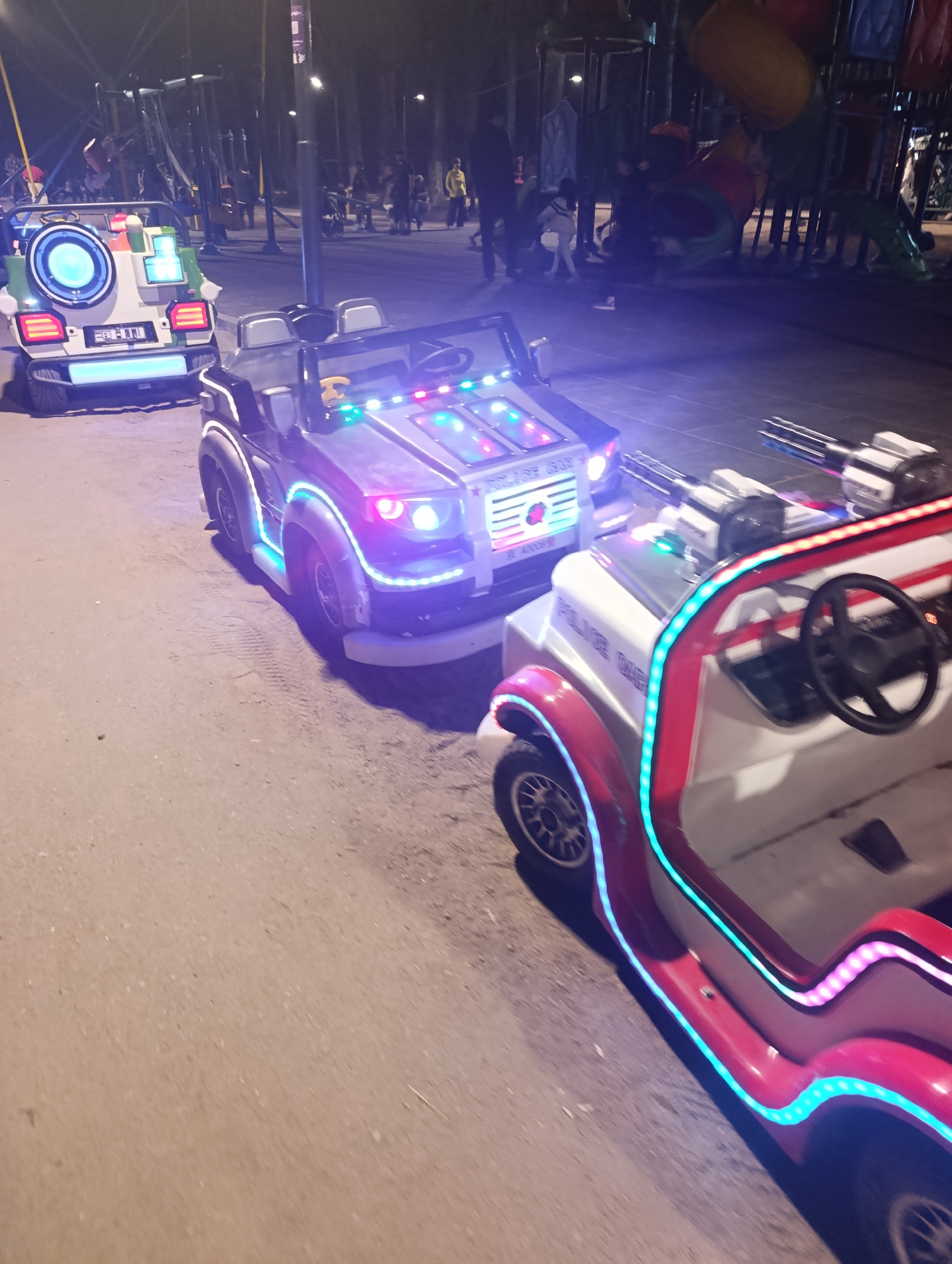 The image depicts a nighttime scene at what appears to be an amusement park or fairground in Bosteri, Kyrgyzstan. In the foreground, two brightly lit, child-sized electric vehicles are positioned on a paved surface. The vehicle on the right is white with red accents and features prominent LED lighting in various colors, including blue and pink. It has a steering wheel and what appear to be mounted toy machine guns. The word "POLICE CAR" is visible on its side. The vehicle on the left is primarily white and green with a large circular illuminated element on its rear and blue and red LED lights. Behind these vehicles, playground equipment, including slides and climbing structures, are visible, illuminated by artificial lighting. Several people, mostly indistinct due to the low light conditions, are present in the background, some standing and others seemingly in motion. The overall atmosphere is one of an outdoor entertainment venue at night.