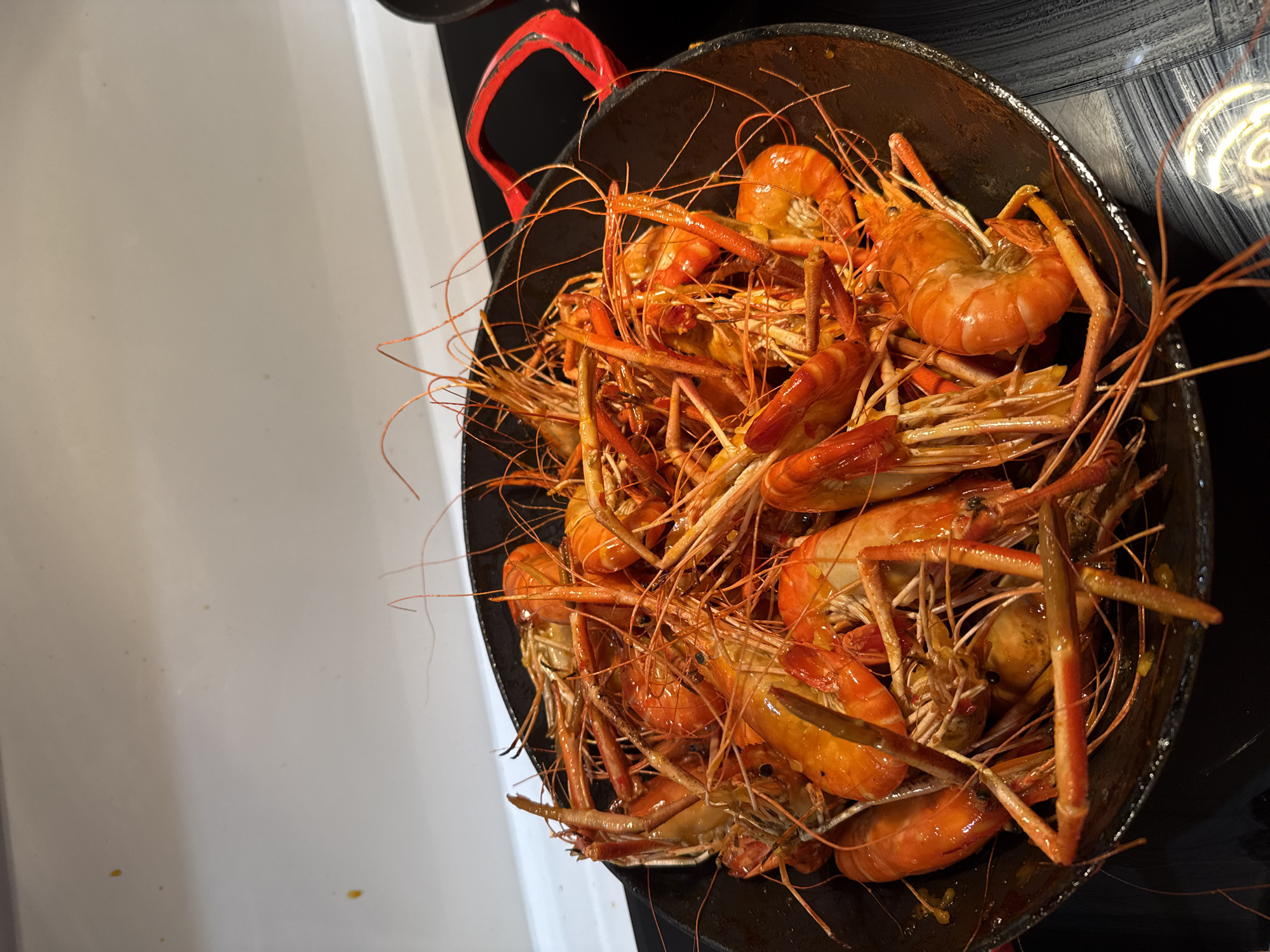 A close-up, overhead shot displays a generous serving of cooked prawns in a dark, round pan with red handles. The prawns are a vibrant orange-red color, indicating they are cooked. Their legs and antennae are long and delicate, some intertwined with each other. The pan appears to be on a black surface, possibly a countertop, with a white background behind it. The lighting suggests an indoor setting, possibly a restaurant or kitchen. There are no people visible in the image, and no specific details about the location in Nha Trang Ward, Vietnam are provided within the visual information.