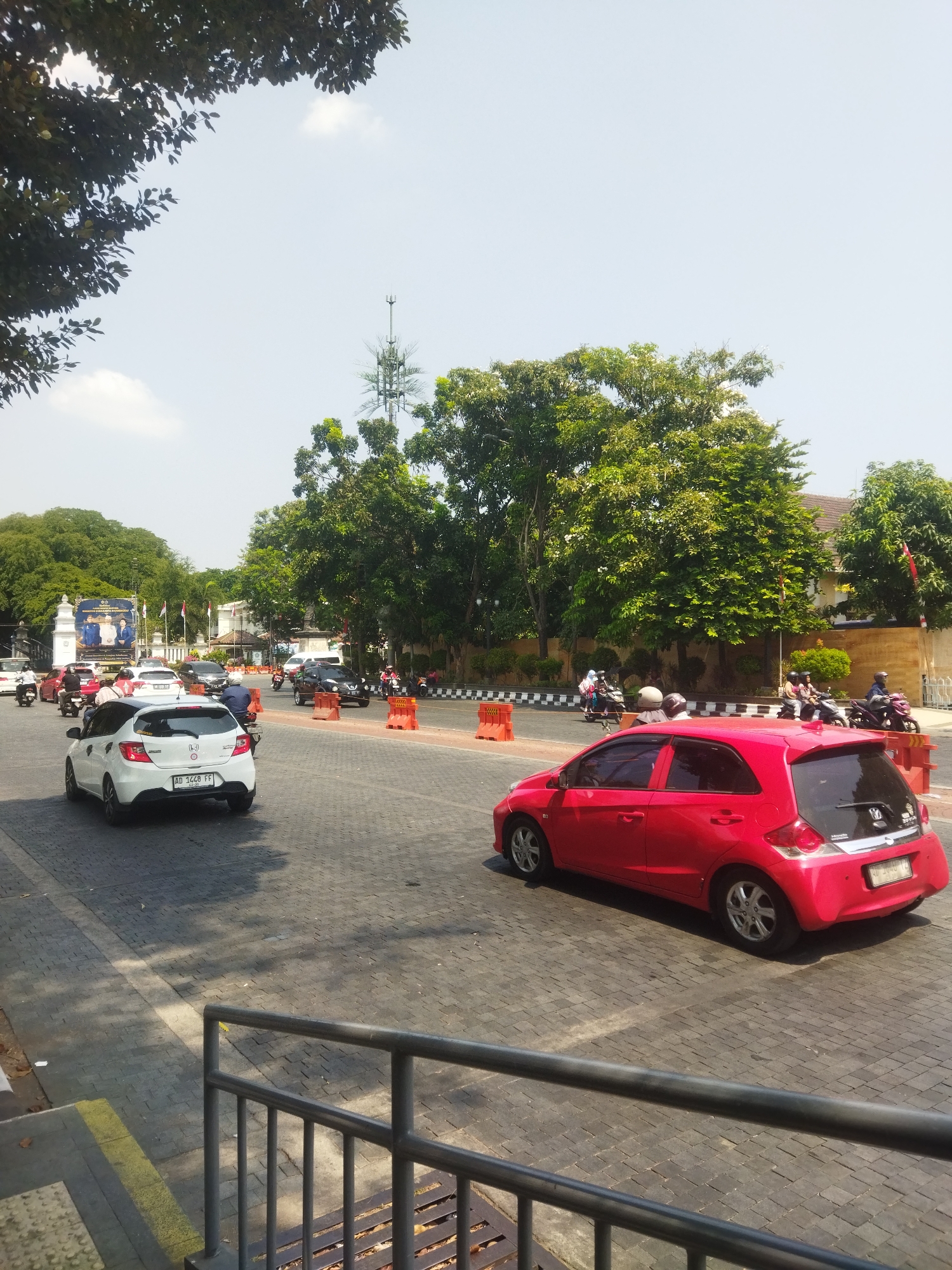 The image shows a street scene in Surakarta, Indonesia, likely on a sunny day given the clear sky and shadows. In the foreground, a red hatchback car is visible from the rear, parked on a paved street with a cobblestone-like pattern. A metal railing and part of a structure are in the lower left of the frame. Further down the street, a white hatchback car is in motion, and several other vehicles, including cars and motorcycles, are also present, indicating moderate traffic.
On the left side of the street, a large banner displaying portraits of two individuals and some text is prominently displayed on a gate-like structure, possibly indicating a government building or event. Trees line the street, providing shade and greenery, and a tall, slender communication tower is visible in the distance against the sky. Orange traffic cones are placed along the road, suggesting ongoing construction or traffic management. The overall environment appears to be an urban setting within Surakarta, characterized by a mix of modern vehicles and traditional pavement.