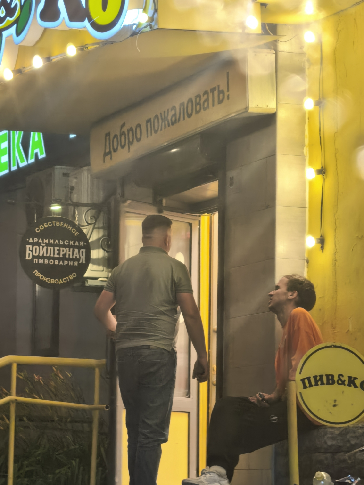 The image shows a street-level view of a building entrance at night. A sign with neon green lettering that says "ЕКА" is visible on the left. Above the entrance, a sign reads "Добро пожаловать!" in black lettering on a light-colored background. Two men are standing near the entrance. One man, wearing a green t-shirt and jeans, is facing away from the camera. The other man, wearing an orange shirt and black pants, is sitting on what appears to be a low wall, looking up and to his right. A circular yellow sign with "ПИВ&К" written on it is visible to the right of the sitting man. The building exterior is yellow, and string lights are hung along the upper edge of the facade. A metal railing, painted yellow, is in the foreground on the left.