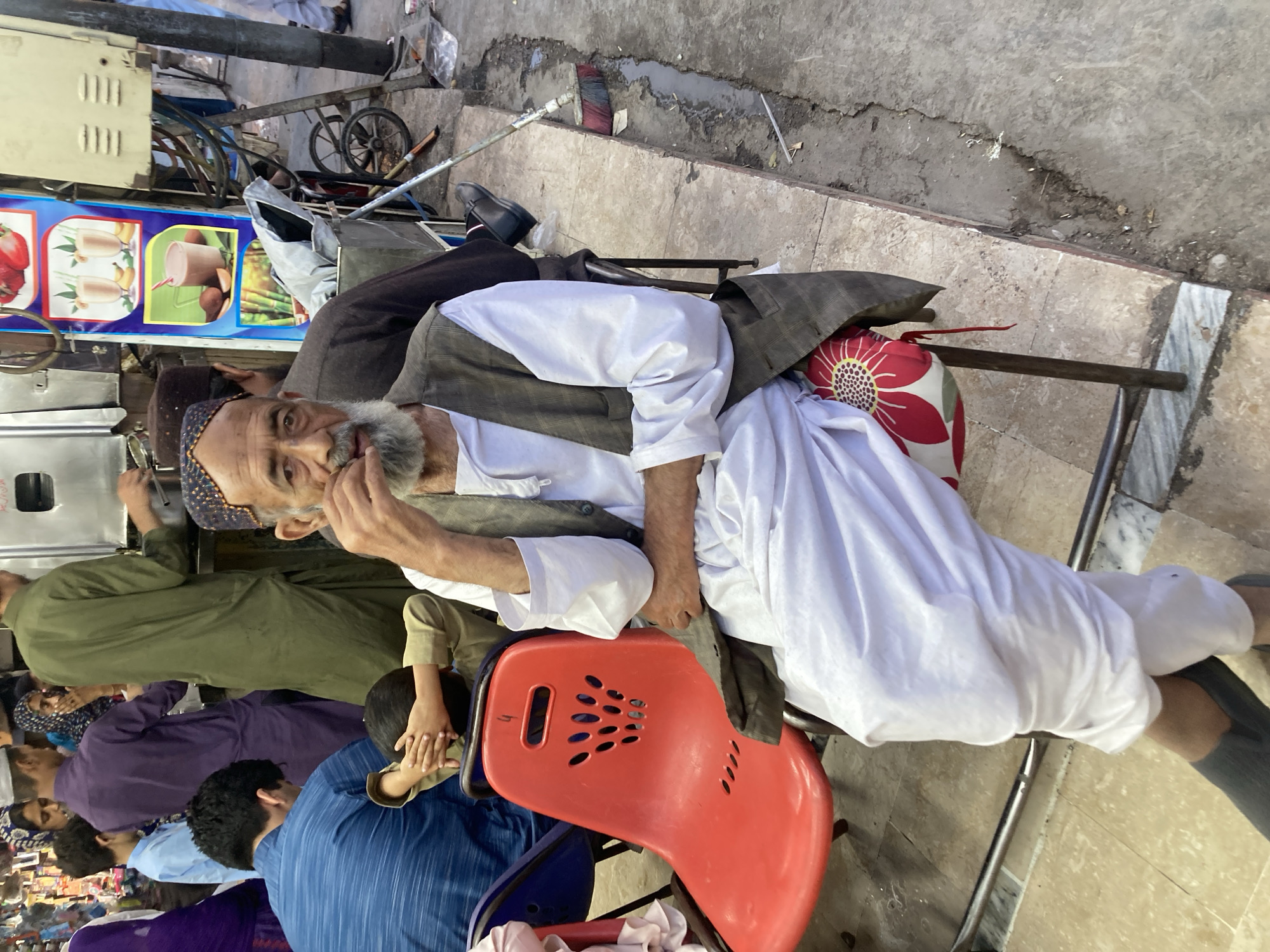 The image shows an elderly man with a grey beard sitting on a red plastic chair on a city street. He is wearing a patterned cap, a white shirt, and a dark vest. He appears to be resting, with one hand near his face. Several other people are standing nearby, some wearing traditional clothing. A juice stand is visible in the background, as well as a cart and other street elements. The location is likely in Quetta City Tehsil, Pakistan, based on the overall appearance and clothing styles of the people.