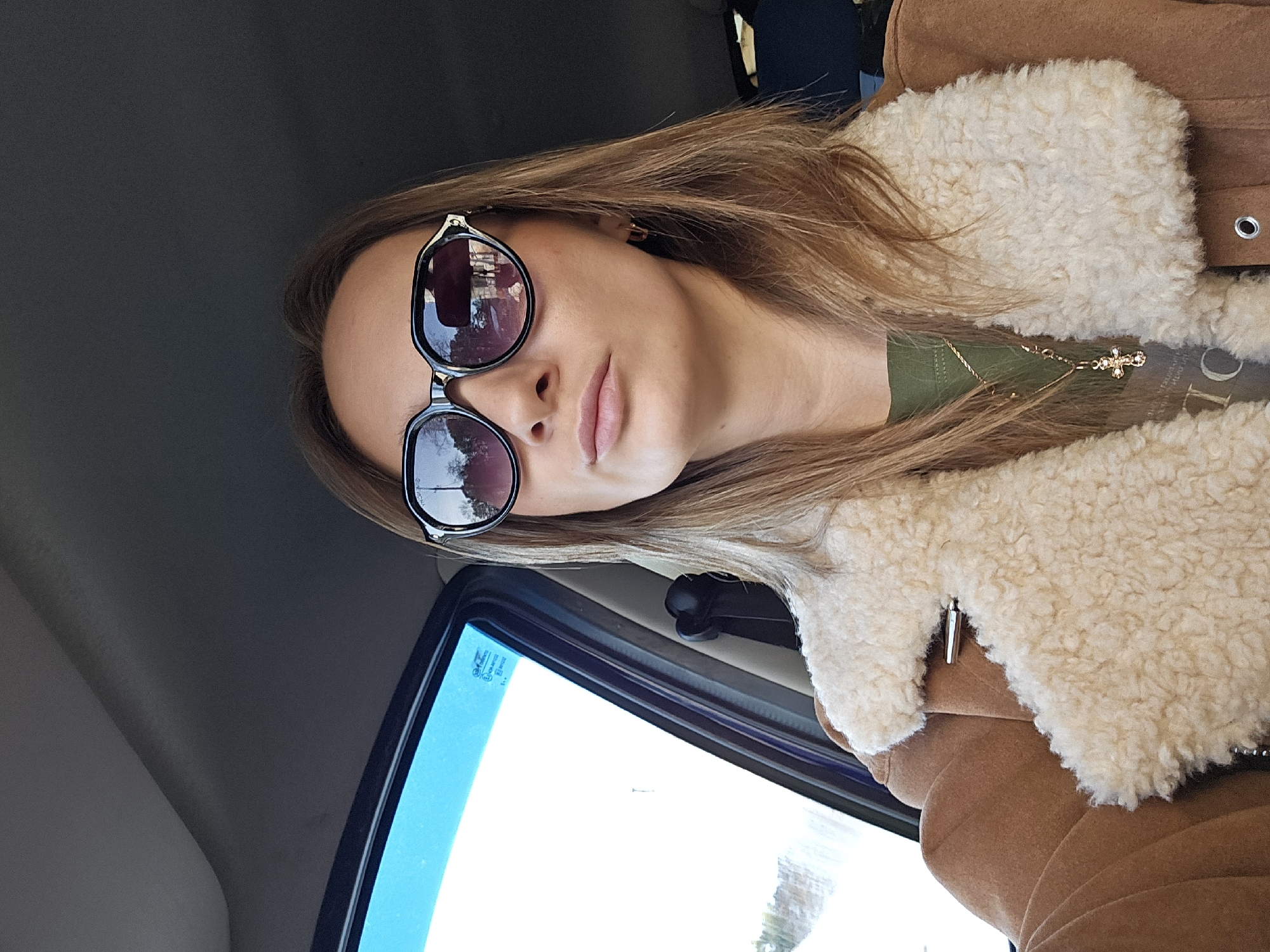 A woman wearing sunglasses, a brown jacket with a white, fluffy collar, and a green shirt sits inside a car. She is also wearing a gold necklace with a cross pendant. The image appears to have been taken inside a vehicle, judging by the window and interior elements. The location is unknown.