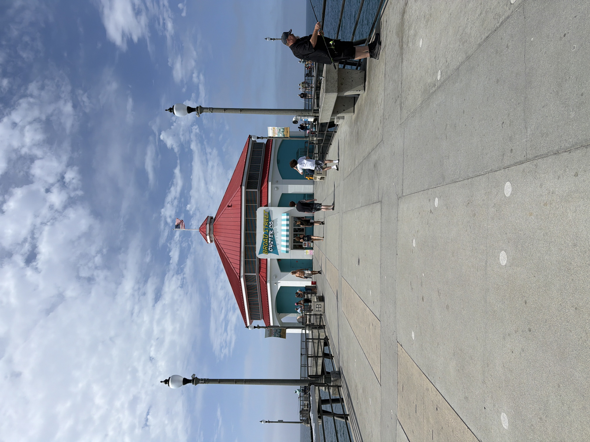 The image depicts the Huntington Beach Pier in Huntington Beach, California, USA. The pier extends into the ocean under a partly cloudy sky. In the foreground, a concrete walkway is visible, with a black metal railing bordering the ocean on the right. Several people are present on the pier: a man is fishing on the right, while others are walking towards or away from a building located further down the pier. This building has a distinctive red, octagonal roof and features signage for "BROAD STREET OYSTER CO." Two lamp posts with white globes stand on either side of the pier's walkway. The ocean is visible to the right and in the distance, with gentle waves. The overall scene is a sunny day on a popular pier.