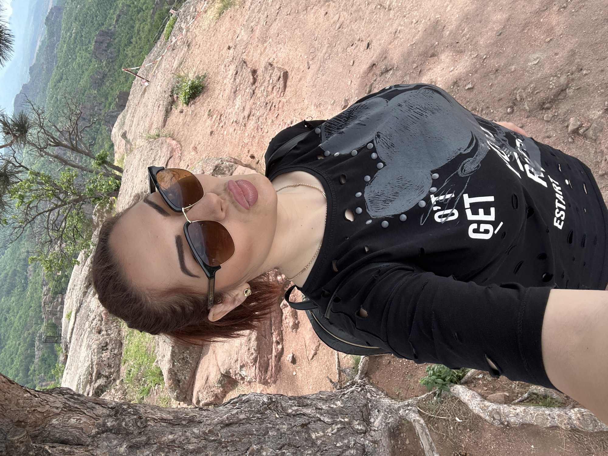 The image is a selfie taken by a woman standing on a rocky outcrop in Belogradchik, Bulgaria. She is wearing sunglasses, a black t-shirt with a graphic design, and a gold necklace. She is pouting slightly for the camera. The background shows a scenic view of the Belogradchik Rocks, with trees and greenery visible in the distance. A tree trunk is visible in the foreground.