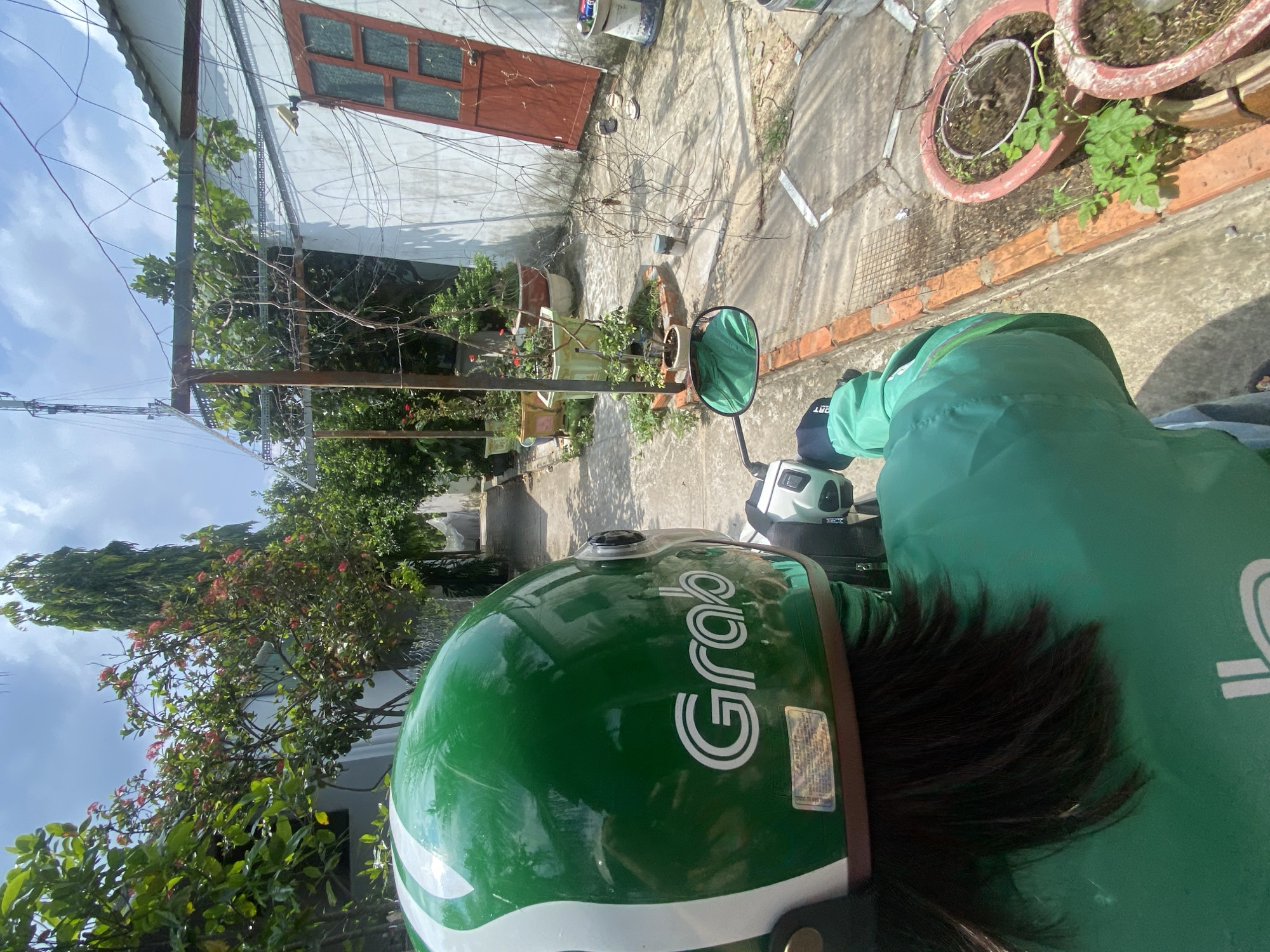 The image shows a person wearing a green Grab helmet and jacket, sitting on a scooter. The scooter is parked on a concrete pathway, with a brick border. In the background, there is a white building with a red door, and a trellis covered in vines. Several potted plants are arranged along the pathway. The scene appears to be in a residential area, with trees and a tall, slender tree visible in the distance. The sky is blue with some clouds. The location is identified as Phu Thuy Ward, Vietnam.