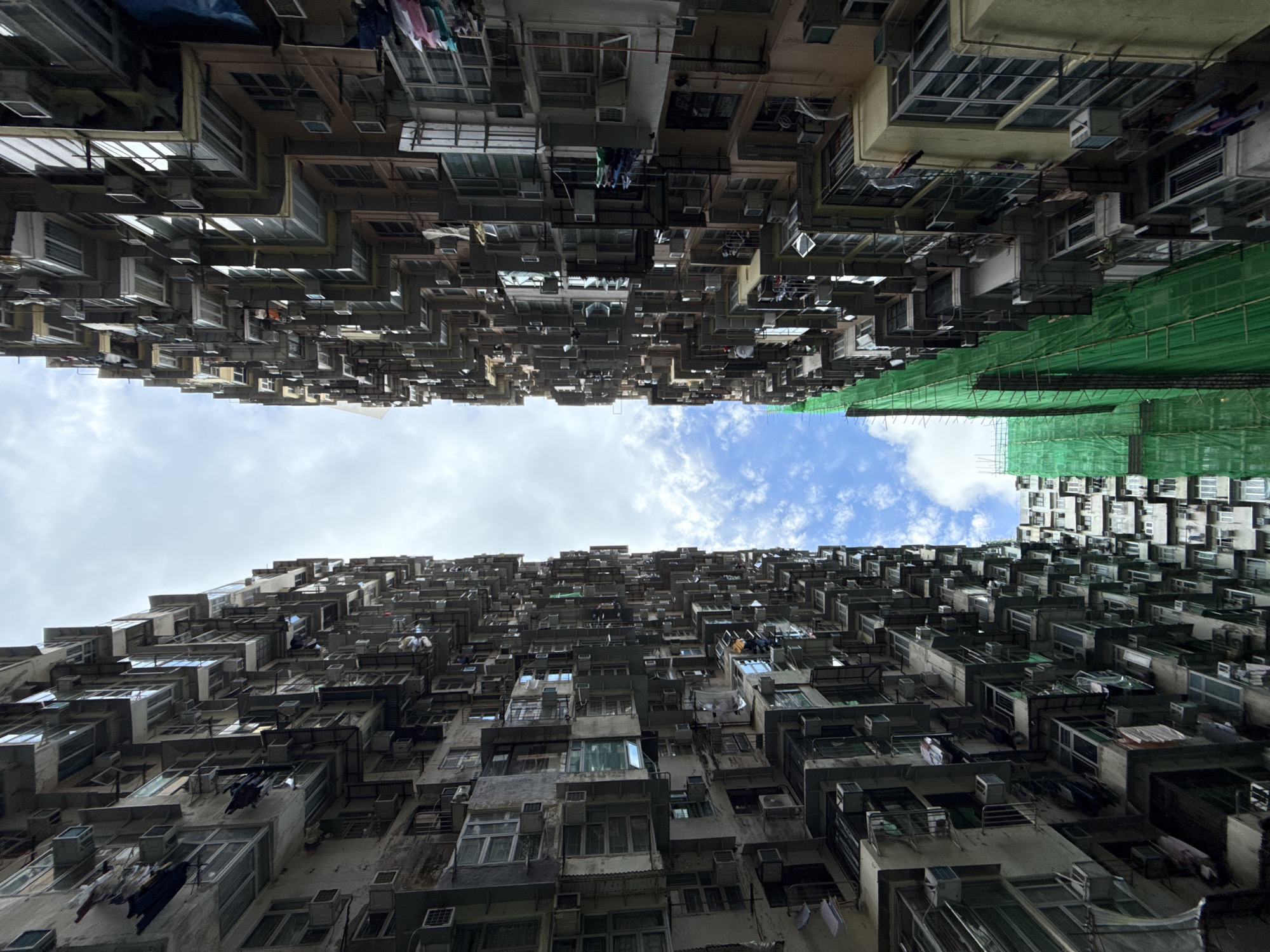 The image captures a low-angle view of a densely packed residential area in Hong Kong, China. The buildings, composed of numerous apartments stacked vertically, dominate the frame. A narrow strip of sky with light clouds is visible between the towering structures. Washing lines with clothes hanging to dry are visible. One of the buildings has construction netting. The architecture is old and weathered, with a monotonous pattern of windows, balconies, and air conditioning units. There are no people visible. The overall impression is one of overcrowding and high-density urban living.
