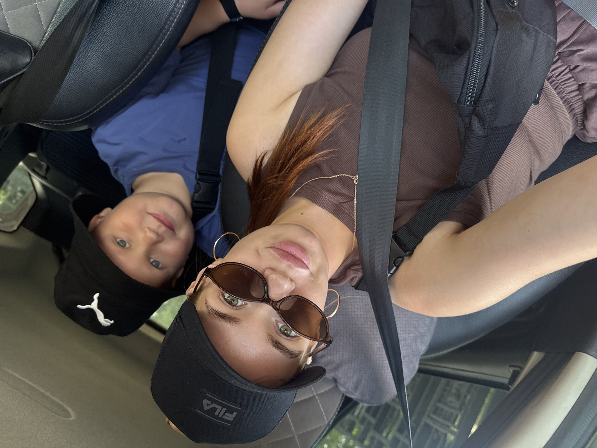 In the city of Chaikovsky, Russia, a woman and a young boy are seated in a car, both wearing hats. The woman, in the foreground, wears a black baseball cap with a "FILA" logo and sunglasses. She is also wearing a brown tank top and has a black fanny pack. The boy, positioned behind her, is wearing a black bucket hat with a white Puma logo and a blue t-shirt. The car's interior is visible, with seat belts buckled across both individuals. The lighting suggests it is daytime, possibly with sunlight filtering into the vehicle.