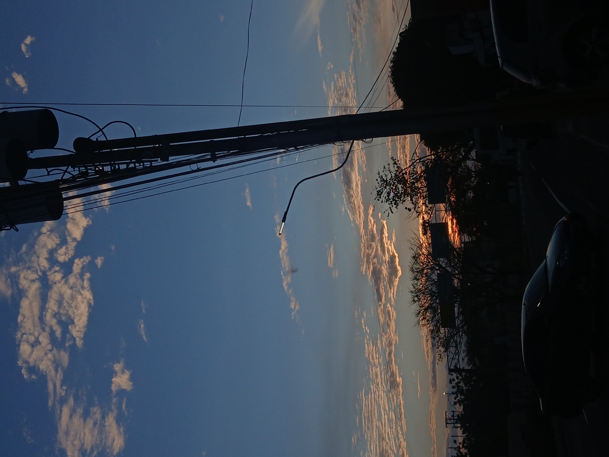 The image shows a street scene in Juan Griego, Venezuela. A utility pole with wires and a street light dominates the left side of the frame, silhouetted against a blue sky with white clouds. The sky transitions from a deeper blue on the left to a lighter blue with orange-tinted clouds on the right. Silhouetted trees and building elements are visible on the right. A dark, indistinct object, likely a vehicle, occupies the far right edge of the frame. The lighting suggests either sunrise or sunset.