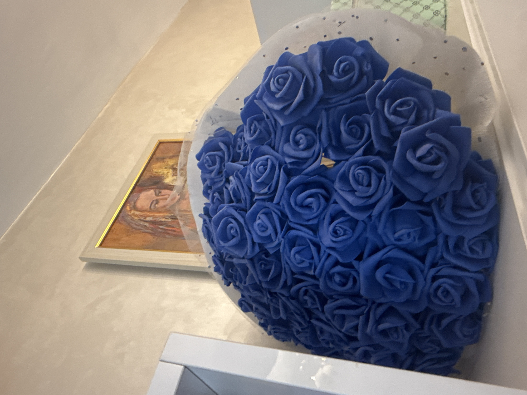 The image displays an indoor scene in Stara Zagora, Bulgaria, centered around a large bouquet of blue roses and a framed picture.
**Subjects:** The main subject is a voluminous bouquet composed of numerous, densely packed blue roses. These appear to be artificial, likely made of foam or a similar material, with defined petals. The bouquet is partially wrapped in a translucent, white netting material adorned with small dark speckles. To the left of the bouquet, a framed picture hangs on the wall. This artwork features a close-up depiction of a woman's face, shown from the chest up, with her eyes looking slightly upwards and to the right. She has brown or reddish-brown hair. The background consists of a plain, light-colored wall, possibly off-white or beige, which exhibits a subtle textured finish. A corner where the wall meets the ceiling is visible in the upper left of the frame. In the lower foreground, a portion of a white surface, likely a shelf or furniture, is visible.
**Activity or event taking place:** No specific activity or event is occurring; the image is a static composition showcasing decorative elements in a room.
**Notable details:** The blue roses are uniformly colored and arranged tightly to form a coherent mass. The white frame of the picture has a thin golden inner trim, highlighting the artwork. The lighting is soft and even, suggesting indoor artificial illumination. The perspective of the photograph is from a slightly low angle, looking upwards at the subjects.
**Visible text:** There is no visible text present in the image.