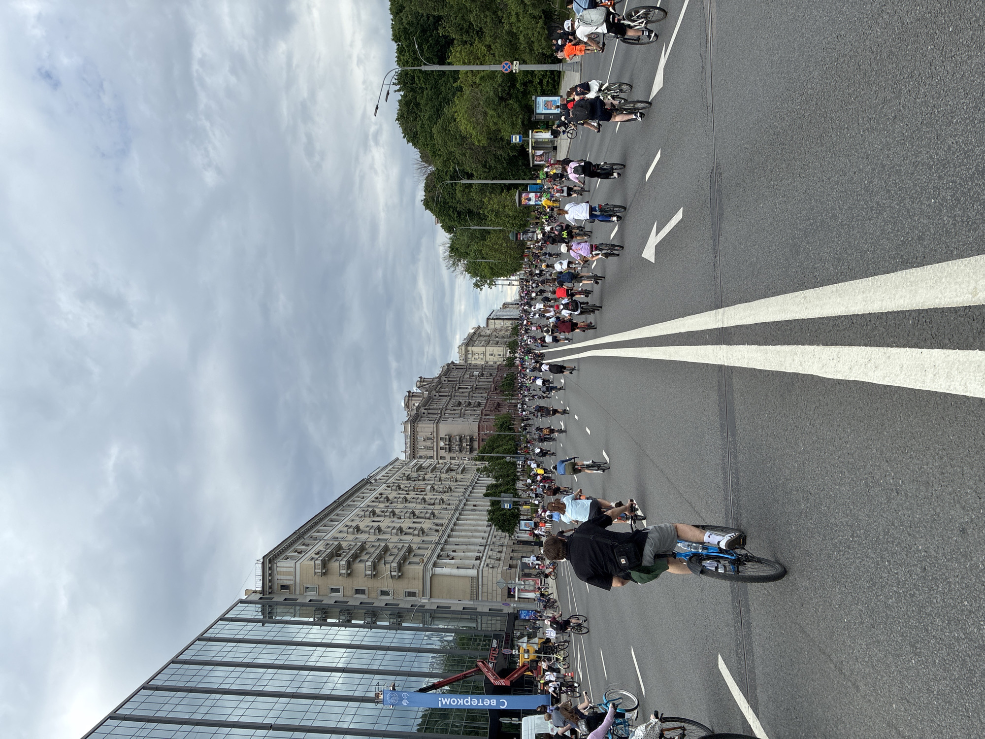 The image shows a large group of cyclists riding down a wide avenue in Moscow, Russia. The street has white lines marking lanes, and a modern glass building is on the left side of the road. A taller, more traditional building with many windows is visible further down the street. Overhead, there are many grey clouds. A banner with Cyrillic text is visible on the left, along with a cherry picker. The cyclists vary in age, attire, and bicycle type.