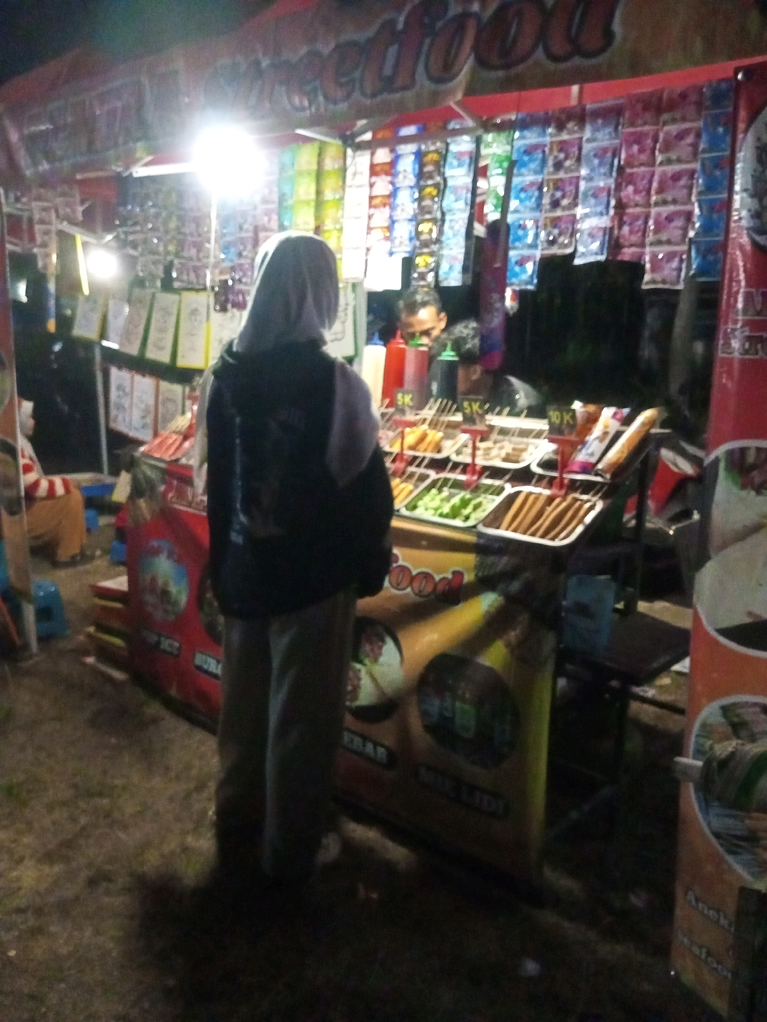 A nighttime outdoor food stall in Ngadikerso, Indonesia, is captured in this image. A person wearing a grey hijab and a dark jacket is standing in the foreground, facing away from the camera and towards the stall. The stall displays various food items on skewers, arranged in trays. Packaged snacks are hung above the food display. In the background, a dimly lit area suggests the presence of other stalls or structures. The ground appears to be covered in dirt and grass.
