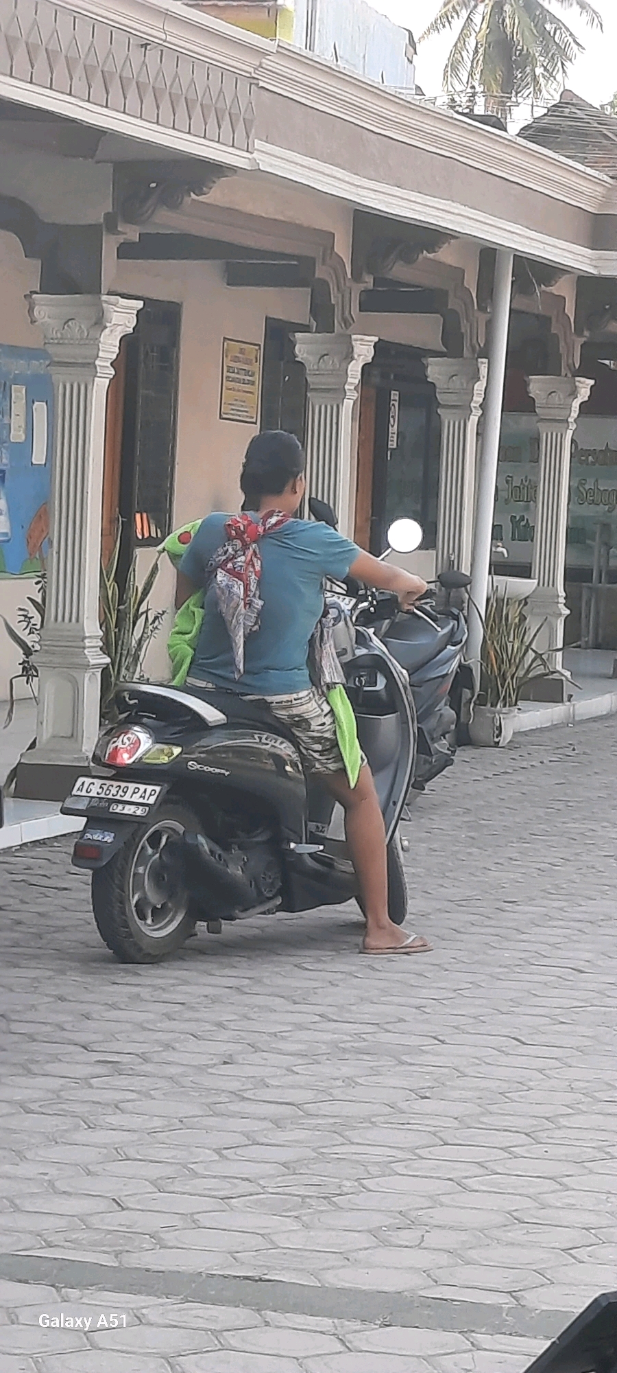In the city of Selopuro, Indonesia, a person is seated on a black scooter, facing away from the camera. The person is wearing a blue t-shirt and patterned shorts. A red and white patterned scarf is tied around their neck and draped over their shoulders. A bright green object, possibly a stuffed toy, is held on their lap. The scooter's license plate reads "AG 5639 PAP". The scooter is parked on a paved surface with hexagonal-shaped stones. In the background, a building with decorative pillars and an awning is visible. Several potted plants are also present in the background. The overall lighting suggests it is daytime.