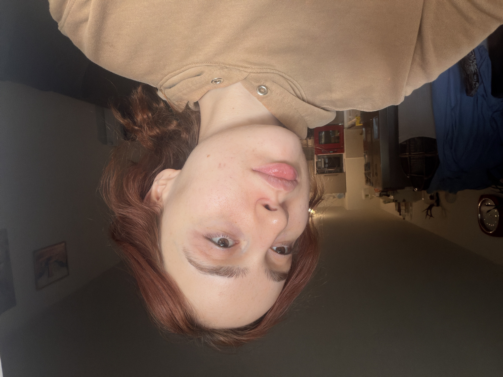 A person with fair skin, auburn hair, and a tan hooded sweatshirt is depicted. The photo is taken from a slightly downward angle, showcasing the individual's face, neck, and a portion of their torso. The background features a dimly lit room, which includes a red microwave and kitchen appliances. There is no indication this image was taken in Strasbourg, France.