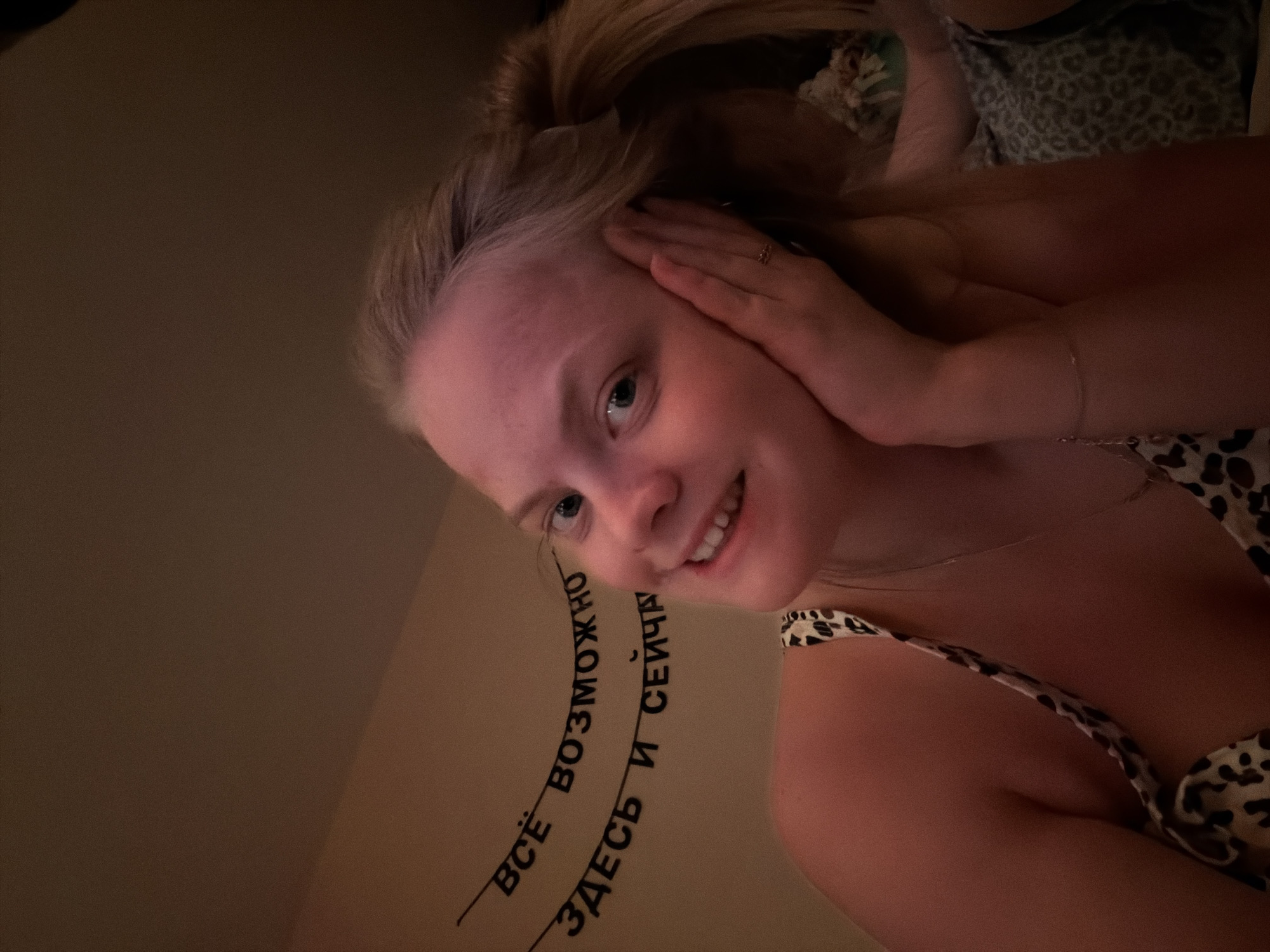 A selfie shows a young woman with light skin and blonde hair pulled back. She is smiling and touching her face with her hands. She is wearing a cheetah-print top. Behind her, on the wall, are the Russian words "ВСЁ ВОЗМОЖНО ЗДЕСЬ И СЕЙЧАС", which translates to "Everything is possible here and now." The location is unknown; there is nothing in the image to suggest it is in Izhevsk, Russia.