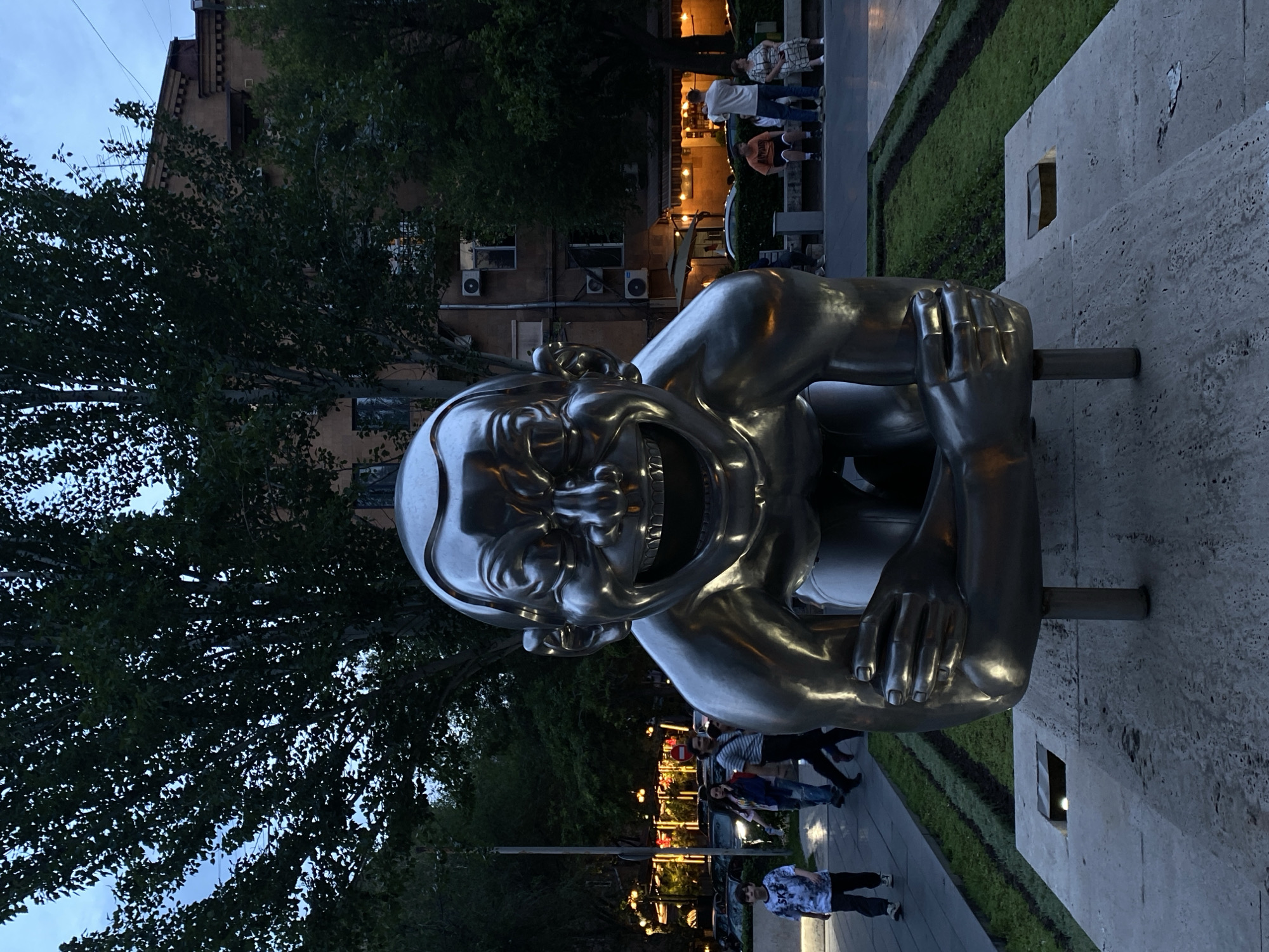 The image presents an evening scene in a public space, featuring a prominent, large metallic sculpture. The central subject is a highly reflective, stylized human figure depicted in an exaggerated state of laughter, with a wide-open mouth, visible teeth, and joyful, wide eyes. The sculpture, likely made of bronze or a similar polished metal, has a rounded body and large hands resting on its knees, and it is mounted on a low, light-colored stone or concrete structure.
The setting appears to be a city park or a public square in Yerevan, Armenia, during dusk or early evening. The sky above is a muted twilight blue. Dense green trees with abundant foliage frame the background, and through them, parts of a multi-story building with windows and air conditioning units can be seen. Along a paved walkway behind and to the right of the statue, several people are present, casually strolling or sitting, enjoying the cool evening air. Artificial lights from street lamps and buildings cast warm glows, illuminating the scene and adding to the serene, social atmosphere. A strip of manicured green lawn borders the walkway. The overall mood is calm and inviting, with the statue's cheerful expression adding a focal point of joy. No discernible text is visible in the image.