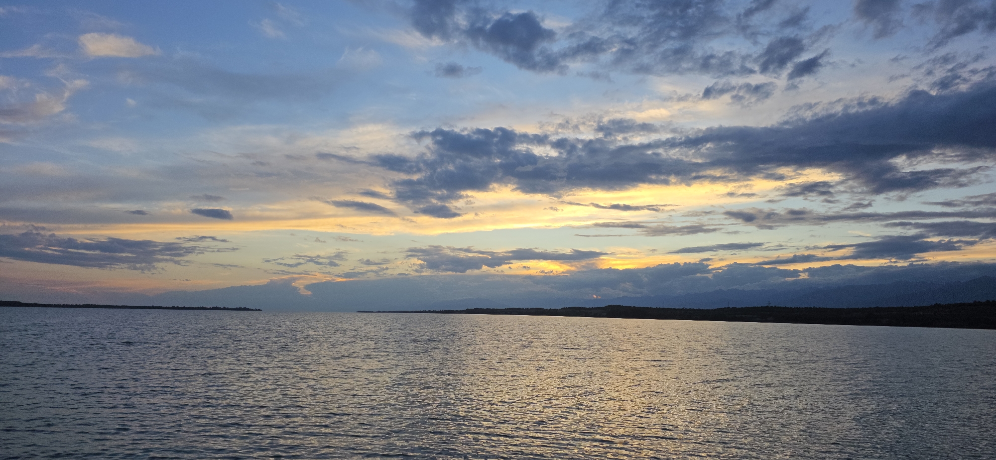 The image shows a wide expanse of water, likely a lake or sea, under a dramatic sunset sky. The water is calm with gentle ripples, reflecting the colors of the sky. The sky is a canvas of blues, grays, and yellows, with clouds forming interesting patterns as the sun sets. A distant shoreline is visible, with a low, dark silhouette of land and mountains against the horizon. The overall atmosphere is serene and picturesque, characteristic of a sunset over a large body of water. Given the location provided, this scene likely depicts Issyk-Kul Lake near the city of Pristan'-Przheval'sk (Karakol), Kyrgyzstan, a popular destination known for its stunning natural beauty and sunsets. There are no people or specific objects clearly identifiable in the image, focusing solely on the natural landscape.