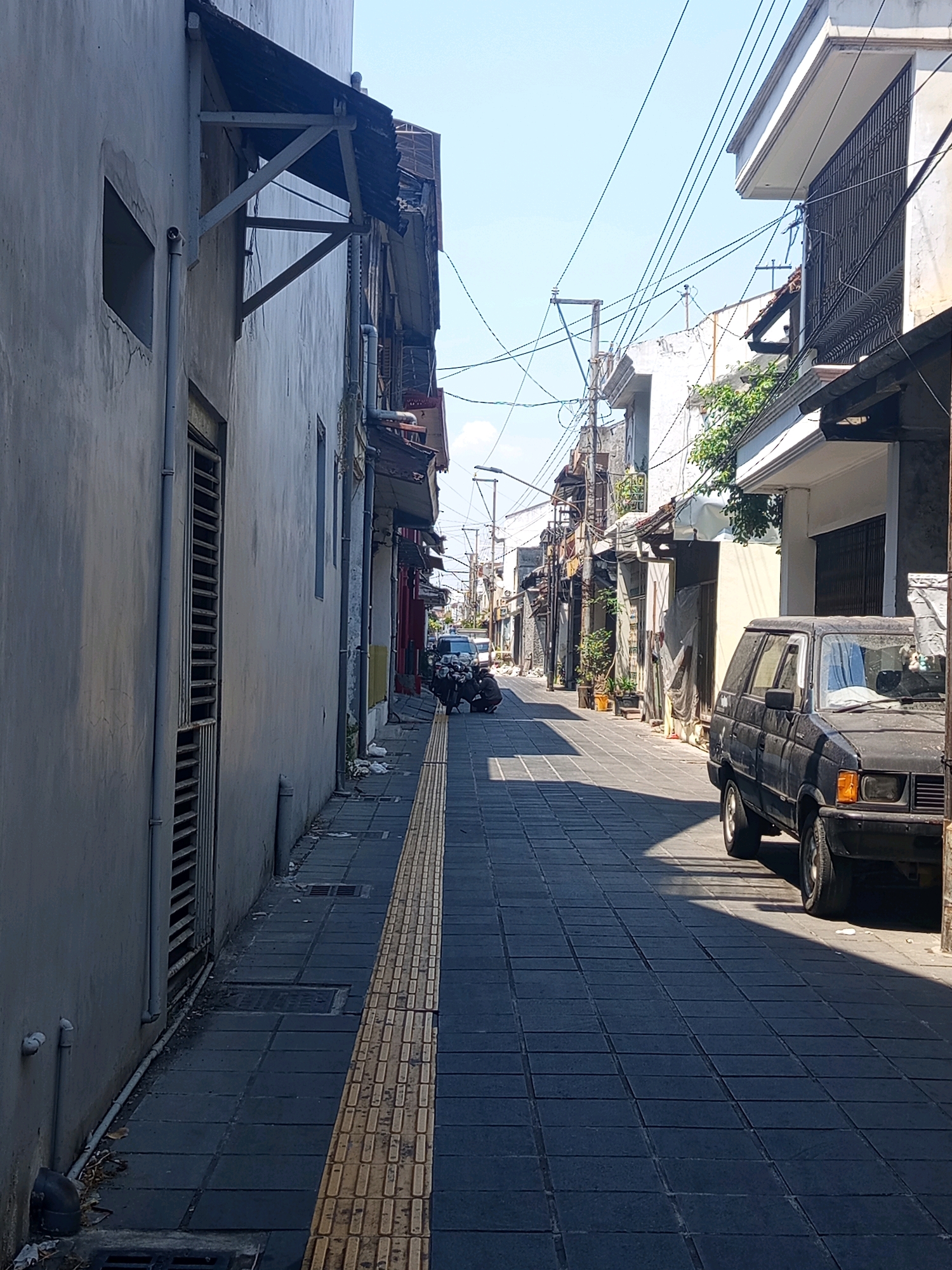 This is a street scene in the city of Semarang, Indonesia. The perspective is from street level, looking down a narrow street lined with buildings on both sides. The street is paved with dark gray square tiles, with a distinct yellow tactile paving strip running along the right side of the sidewalk.
On the left, a light gray building with a slightly textured facade dominates the foreground. It features a metal grate over a vertical opening and a downward-sloping pipe on the lower left. Above this building, an awning made of corrugated metal and supported by angled wooden beams extends outwards.
On the right, a black station wagon is parked parallel to the sidewalk. The car appears to be an older model, with visible wear. In front of the car, a portion of the sidewalk is in shadow. Further down the street on the right, more buildings are visible, some with multiple stories, balconies with iron railings, and various awnings and pipes. Greenery, possibly potted plants, can be seen near some of the buildings.
Several power lines crisscross above the street, supported by poles and extending between buildings. In the distance, more buildings and utility poles are visible, suggesting a densely populated urban environment. A few other vehicles, including motorcycles and possibly another car, are further down the street, along with some people. One person is crouched down near a motorcycle on the left side of the street in the mid-ground. The sky is clear and bright blue. The overall impression is of a busy, lived-in street in a tropical climate.