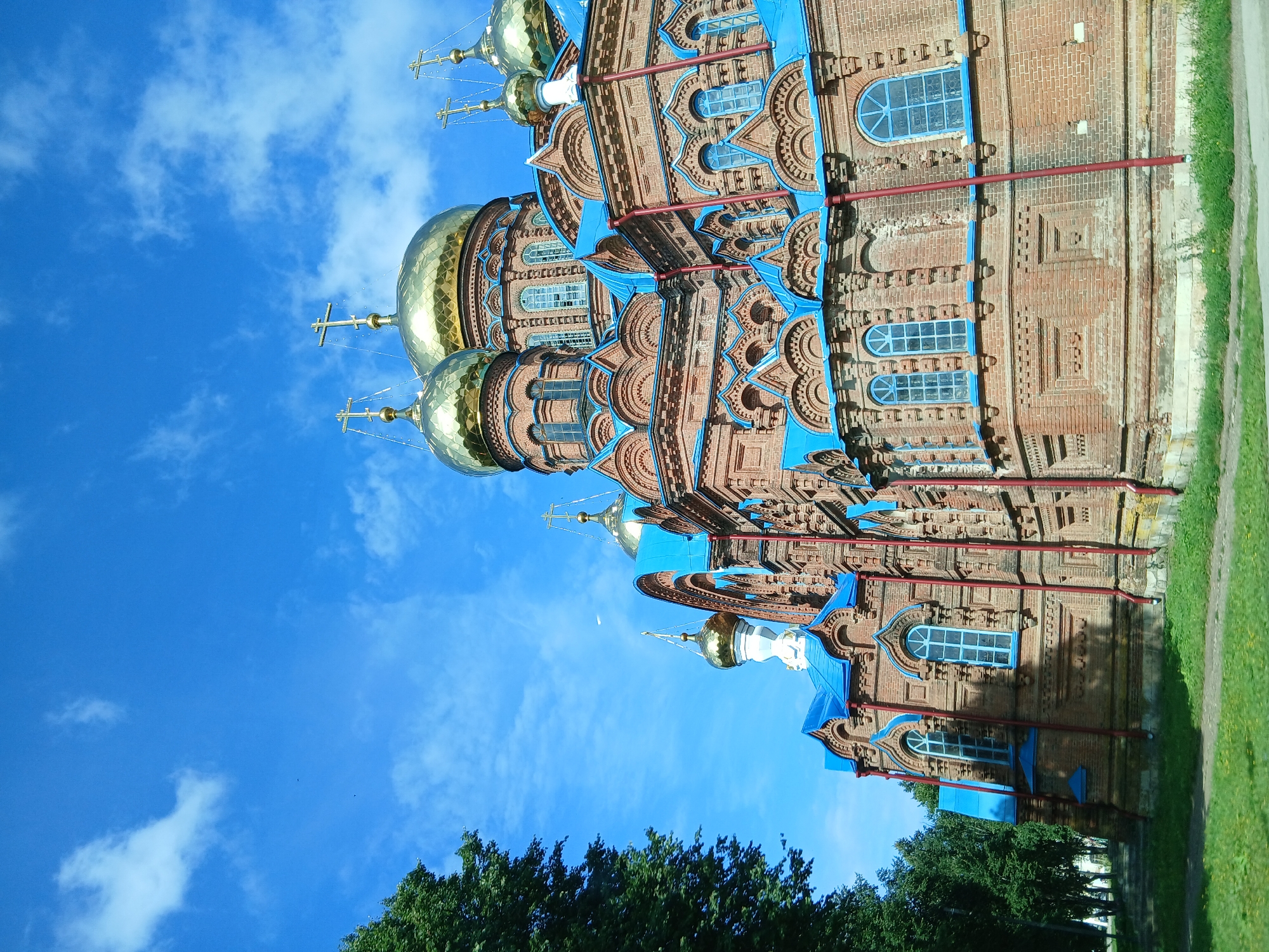 This is a vertical, eye-level, medium shot of the Church of the Resurrection of Christ in the city of Osa, Russia. The red brick church has multiple golden domes with crosses on top, and bright blue roofs and trim. The church is ornate with intricate brickwork detailing. Sunlight illuminates the building, casting shadows. The church is surrounded by green grass and trees on the left side of the frame. The sky is a bright blue with scattered white clouds. No people are visible in the image.