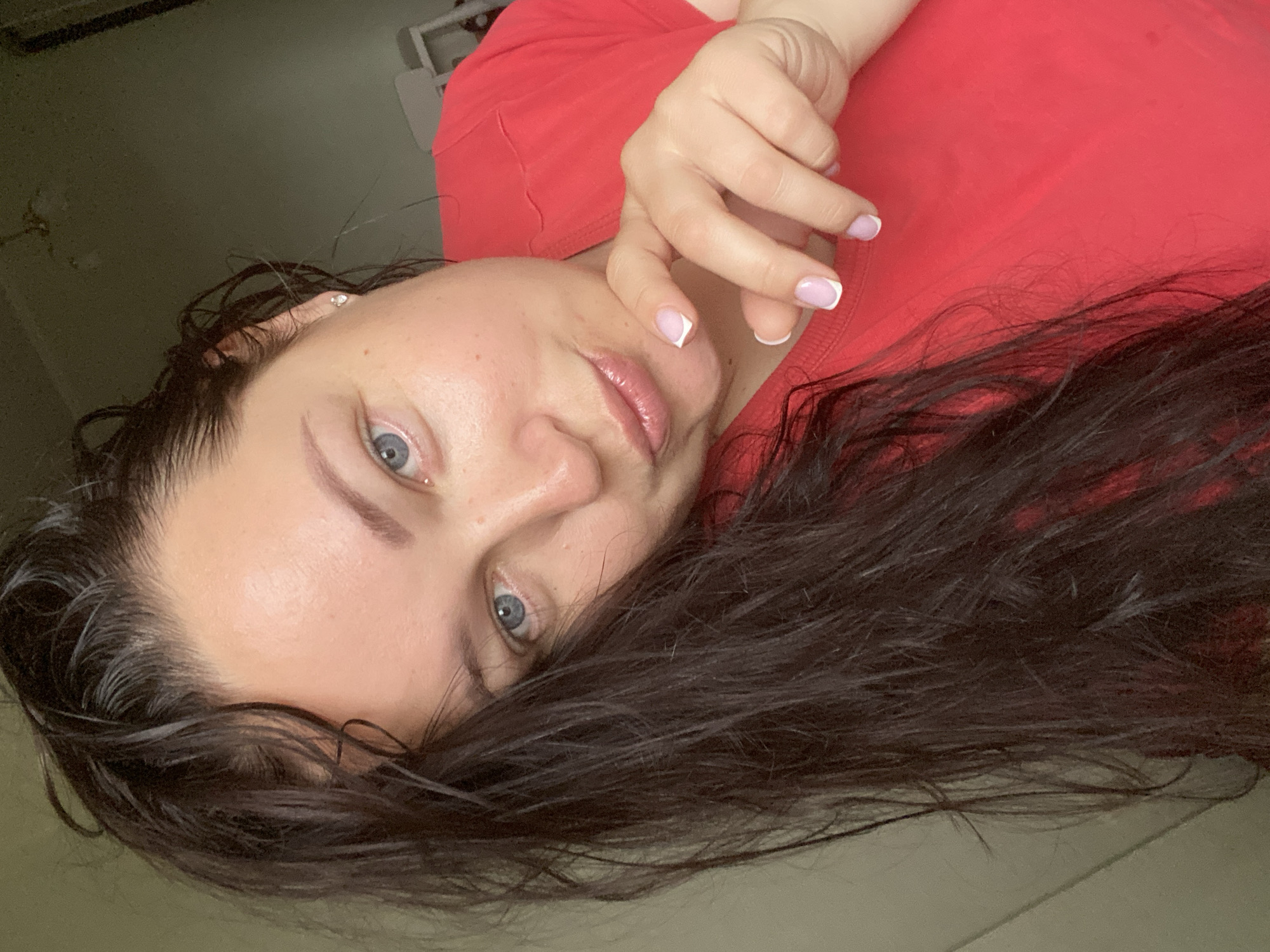A medium close-up shot features a woman with fair skin, blue eyes, and dark, wet hair. She is wearing a red t-shirt. Her fingers with French manicured nails rest near her face. An earring is visible in her left ear. The background is a neutral, out-of-focus wall. There is no indication this photo was taken in Sochi, Russia.