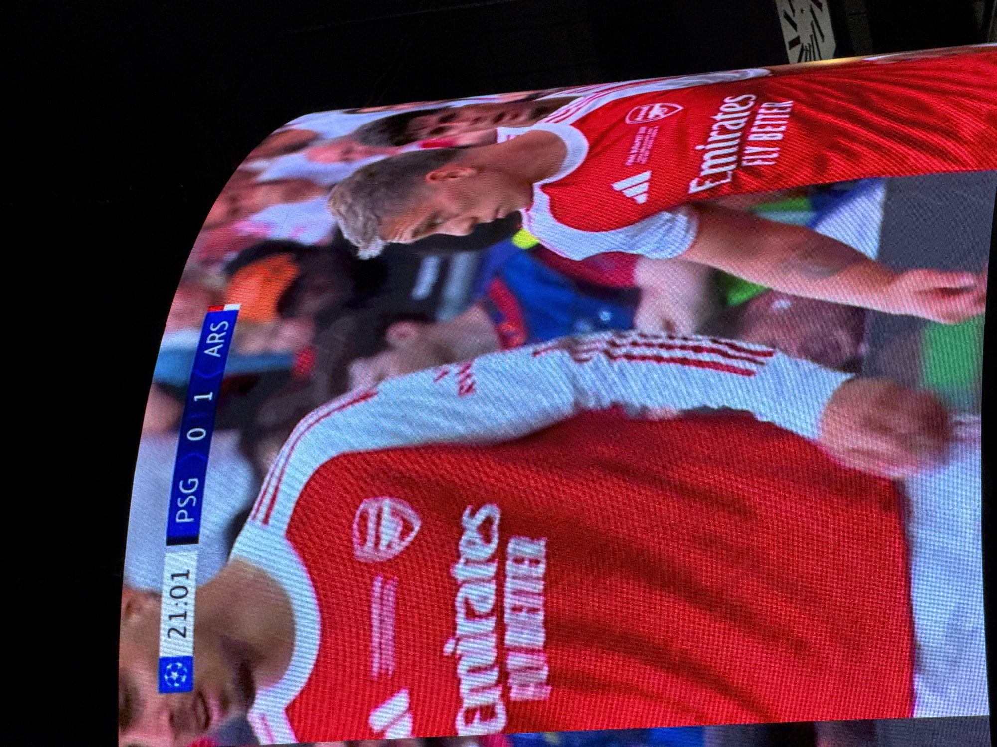 The image shows a television screen displaying a football (soccer) match. The score is PSG 0, ARS 1. The time is 21:01. Two soccer players wearing red and white Arsenal jerseys with the "Emirates FLY BETTER" logo are visible.