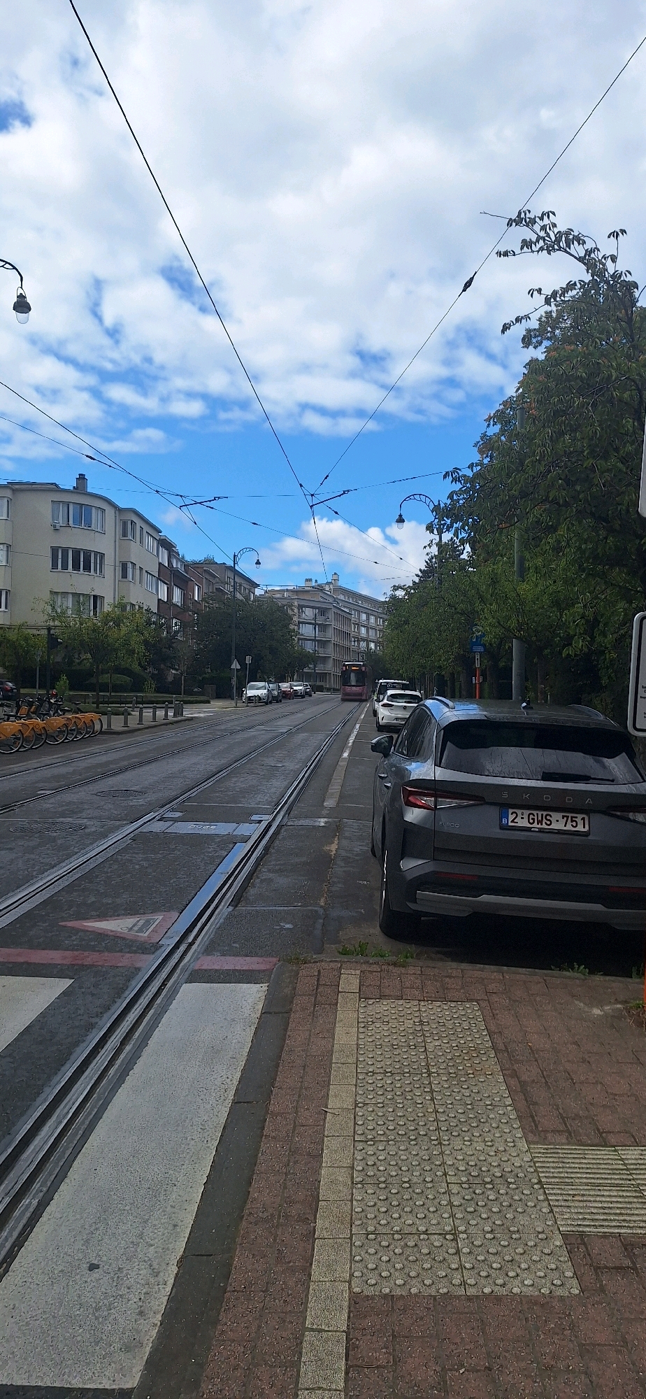 The image shows a street scene in Ixelles, Belgium, with tram tracks running down the middle of the road. A dark gray Skoda SUV is parked on the right side of the street, displaying a Belgian license plate reading "2-GWS-751". Several other cars are also visible, parked along the street and moving in the distance. On the left side of the street, a row of orange bicycles are parked at a bike rack. The buildings lining the street are multi-story apartment buildings, some with balconies. The sky is blue with scattered white clouds, and overhead wires for the tram system crisscross the scene. Trees with green foliage are present on both sides of the street. A pedestrian crossing is marked on the road in front of the parked SUV.