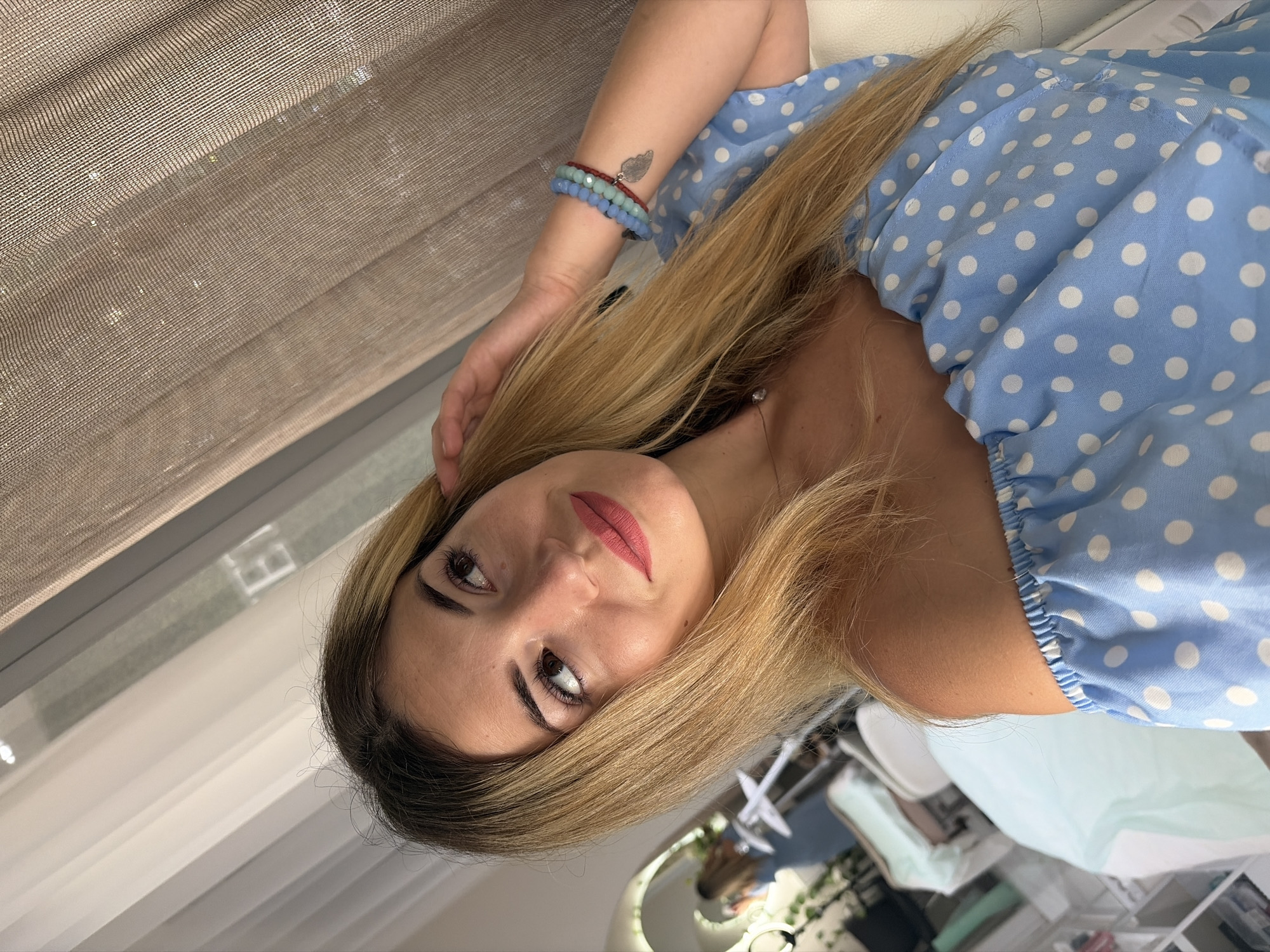 The media file shows a close-up, portrait-style image of a young woman with blonde hair, wearing a light blue dress with white polka dots. She is looking slightly off-camera with a neutral expression. Her left arm is resting on what appears to be a white surface, and she is wearing a bracelet with blue and green beads and a small silver charm. The background is blurred, but suggests an indoor setting with diffused lighting. Part of a window blind or curtain is visible on the left side of the frame, and the overall lighting suggests daytime. There is no visible text in the image.