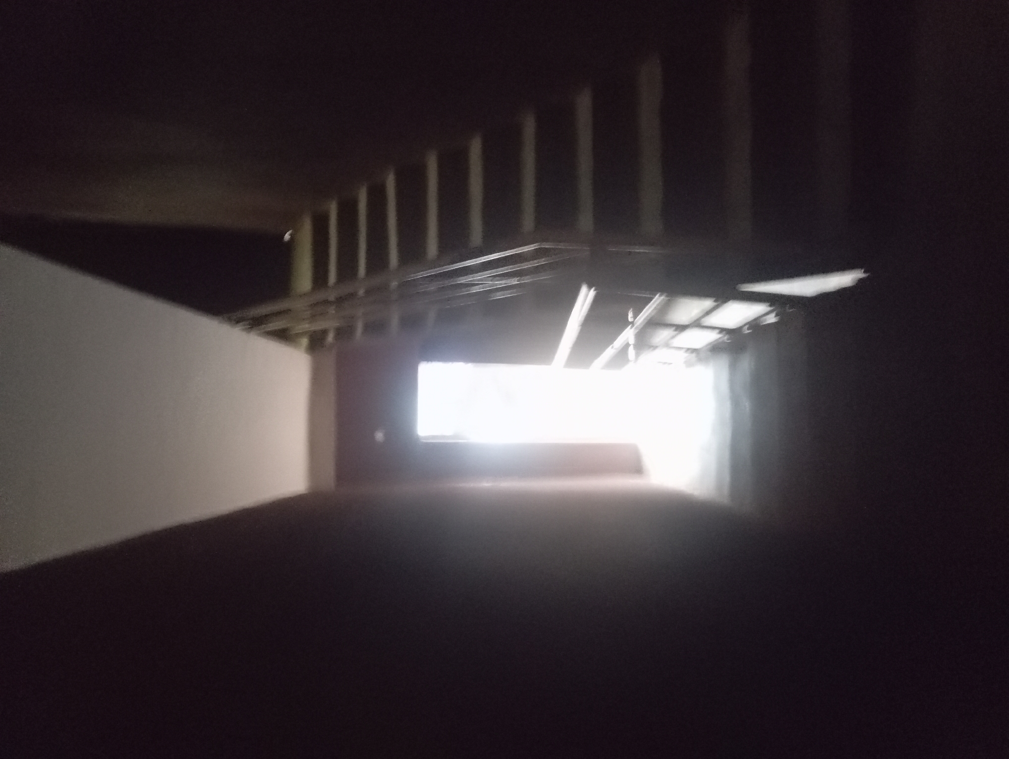 The image depicts a dimly lit, enclosed space resembling a tunnel or a covered walkway, possibly in Nkpor, Nigeria. The space is characterized by a white wall on the left and a dark, indeterminate area on the right. The ceiling has exposed structural elements, including beams and what appears to be metal framework. At the end of the tunnel, a bright light source is visible, obscuring details in that area. The floor is dark and indistinct. No people are visible.
