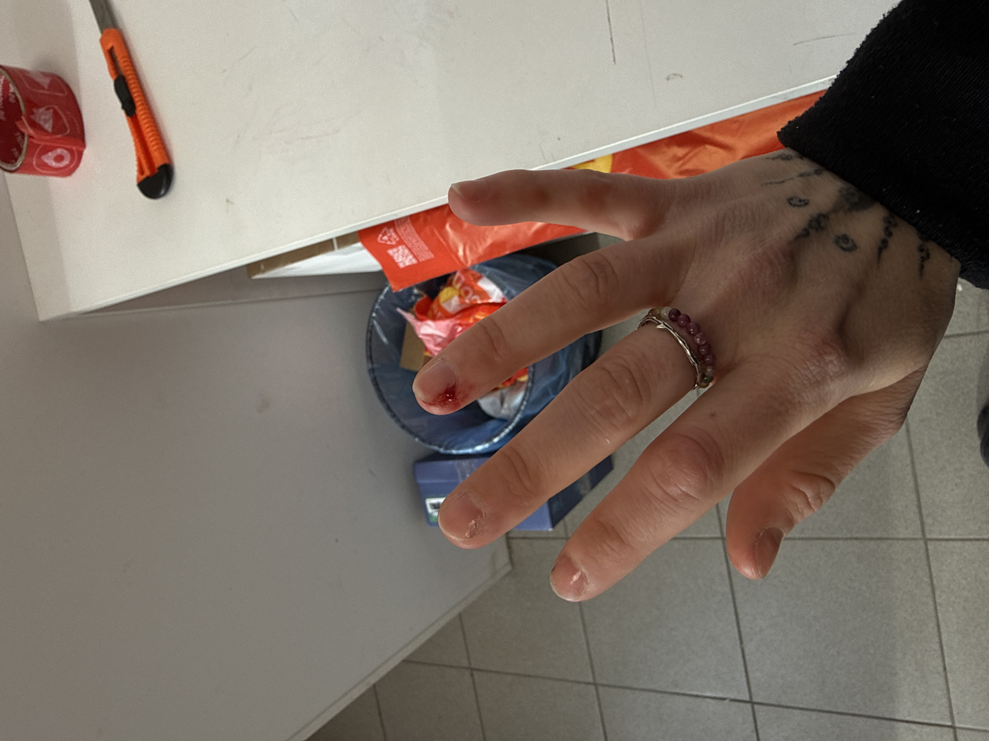 A hand is prominently displayed, showing a small cut on the tip of the index finger. The hand also has a ring on the ring finger and tattoos near the wrist. A utility knife and tape roll rest on a white surface in the background. Underneath the white surface, there's a trash bin with packaging material inside. The floor consists of square tiles. It is not possible to tell the exact location in the city of Saint Petersburg, Russia.